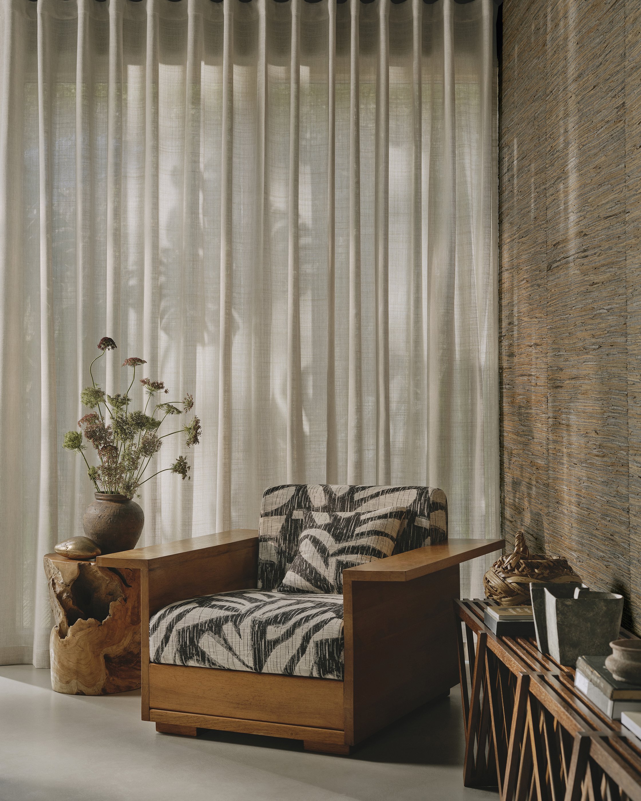 Cozy sitting area with a patterned armchair, a wooden side table, a vase with flowers, and a bamboo screen with beige curtains in the background.