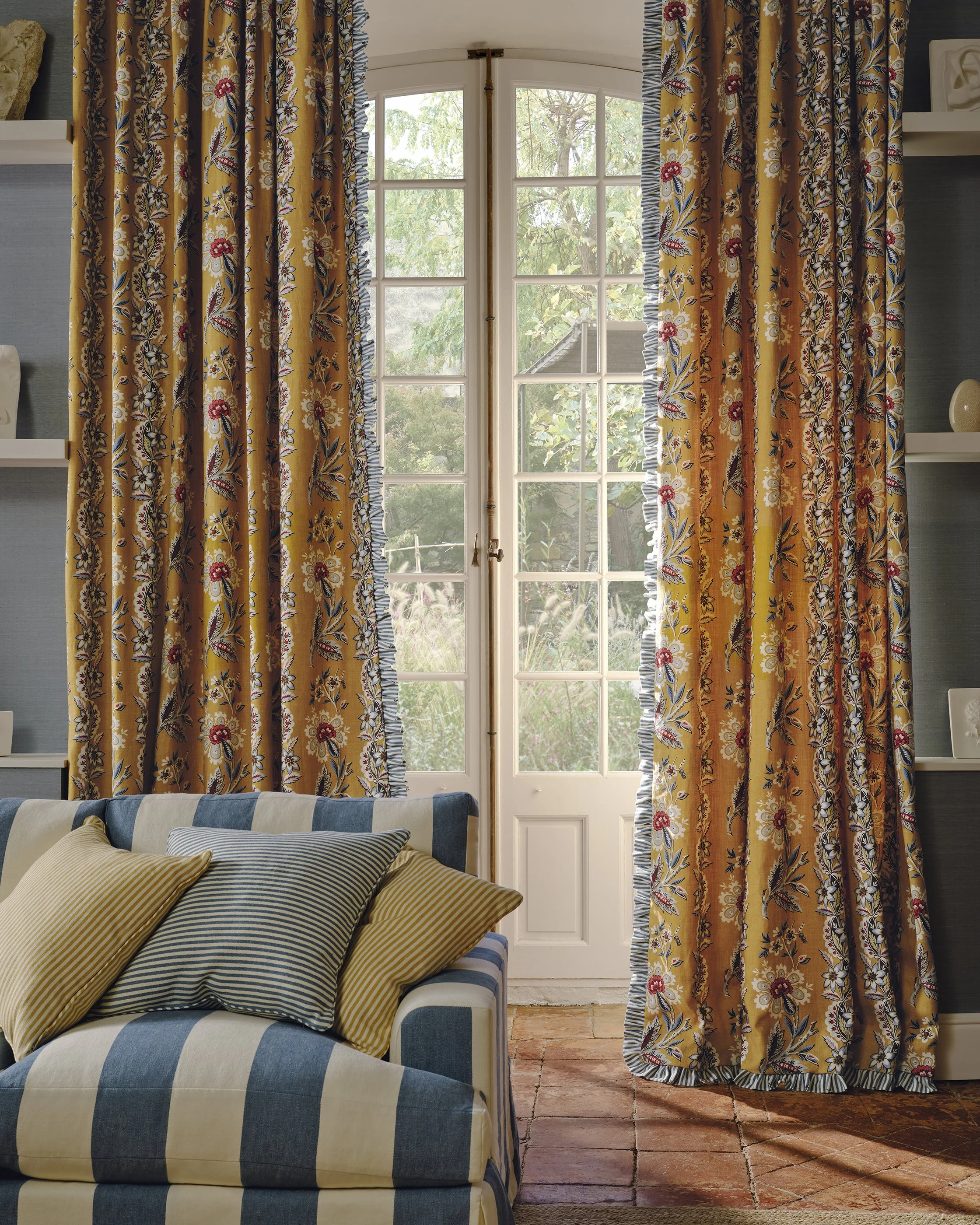 A living room with yellow and floral patterned curtains on a window door, a blue and white striped sofa with yellow and striped pillows, brick flooring, and built-in shelves with decorative items.