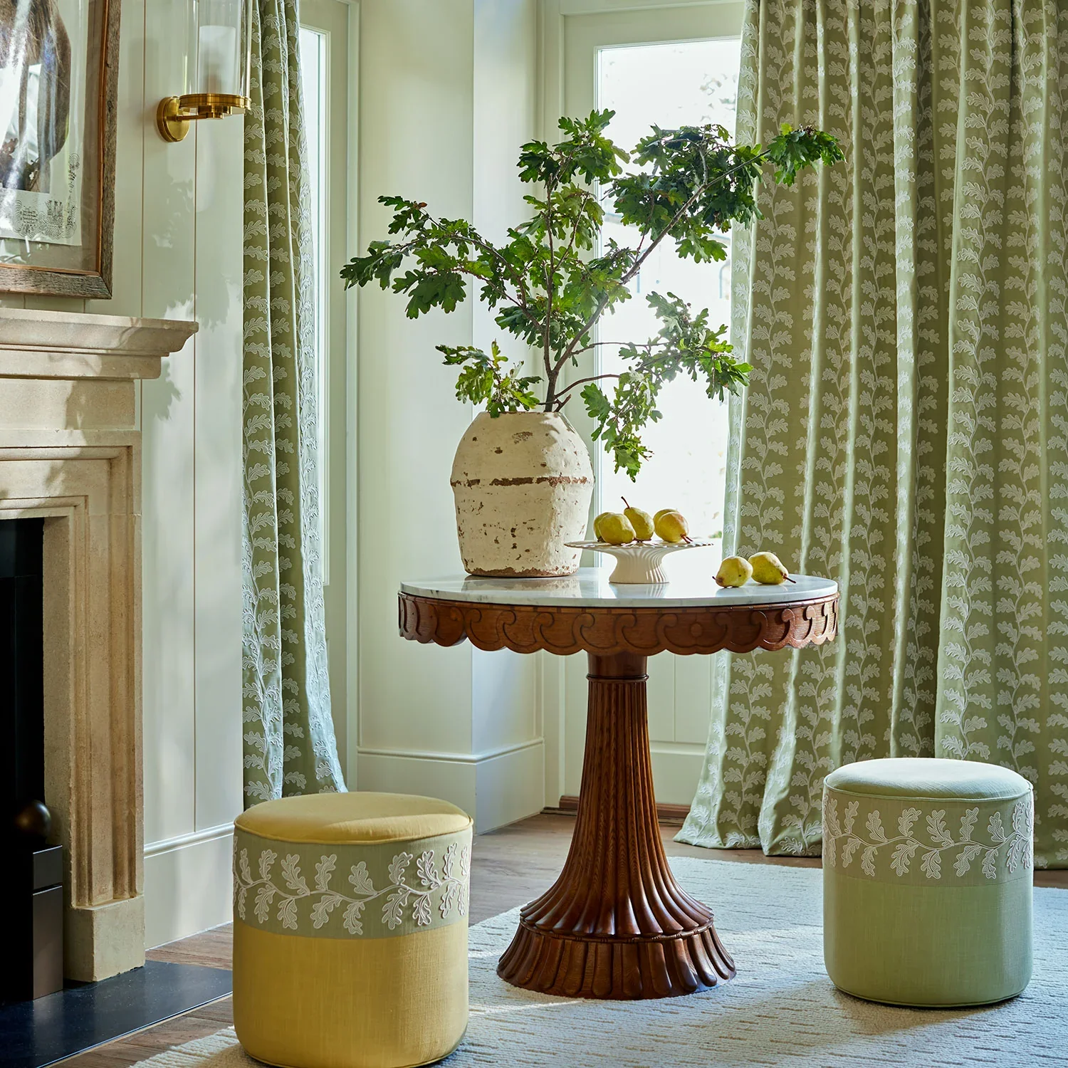 A cozy living room corner with a round wooden table holding a large pottery vase with green leafy branches and a small plate of pears, flanked by two ottomans with yellow and green upholstery featuring white embroidered leaf patterns. Light from a window illuminates the scene, with patterned curtains and a fireplace mantel nearby.