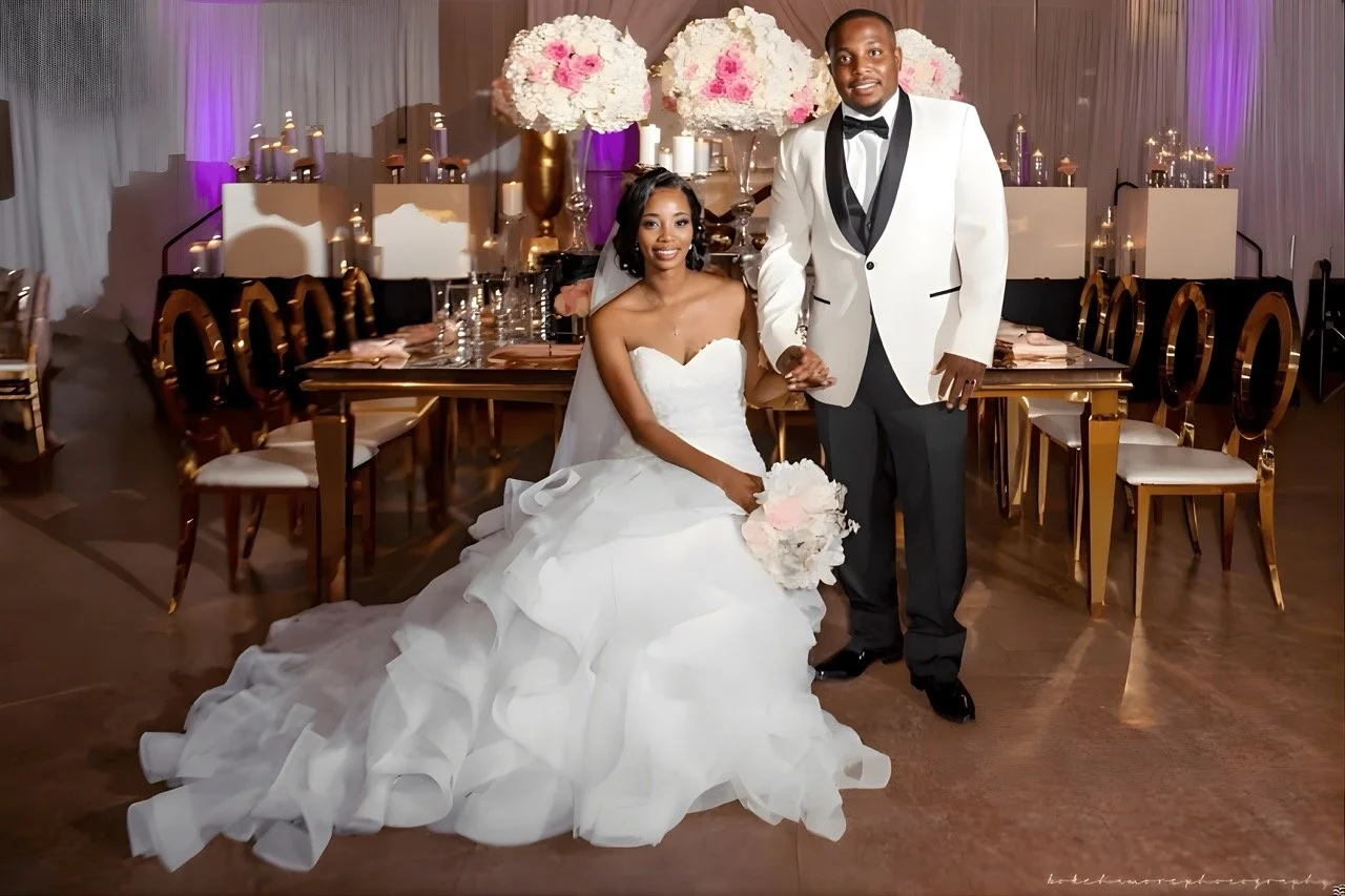 Bride and groom posing together in formal attire in front of an elegant wedding reception table with candles, florals, and gold chairs
