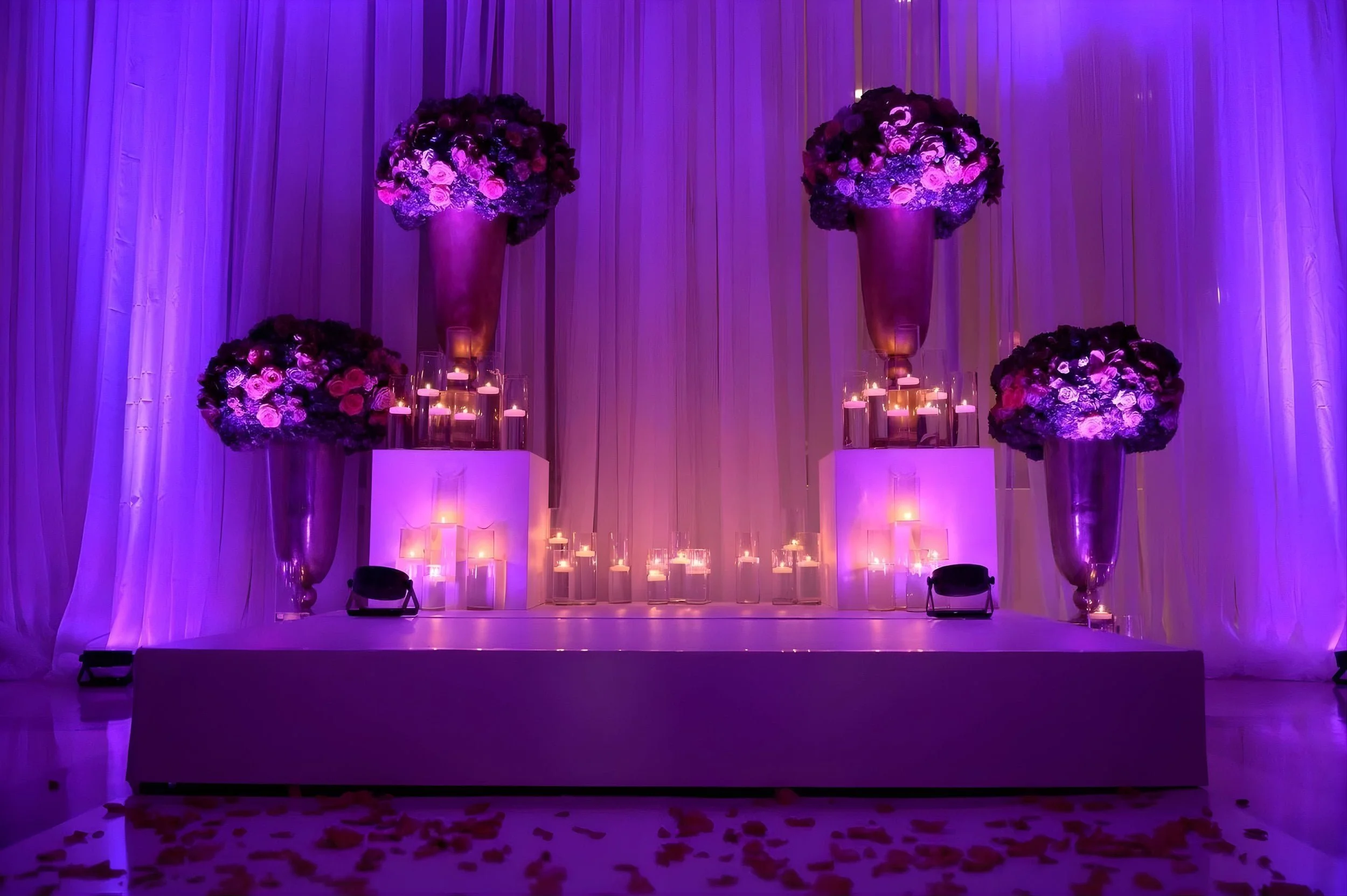 Wedding stage decorated with tall floral arrangements, candles, and draped fabric under purple lighting.