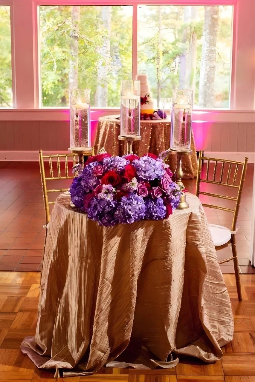 Sweetheart table styled with gold linens, tall candles, and a purple and pink floral arrangement.