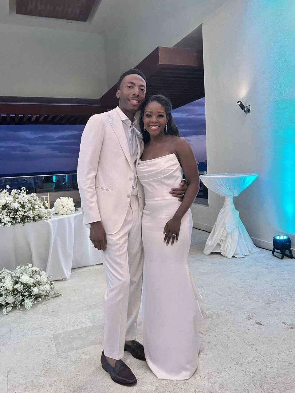 Bride and groom standing together at their wedding reception, dressed in formal attire with floral décor visible in the background.
