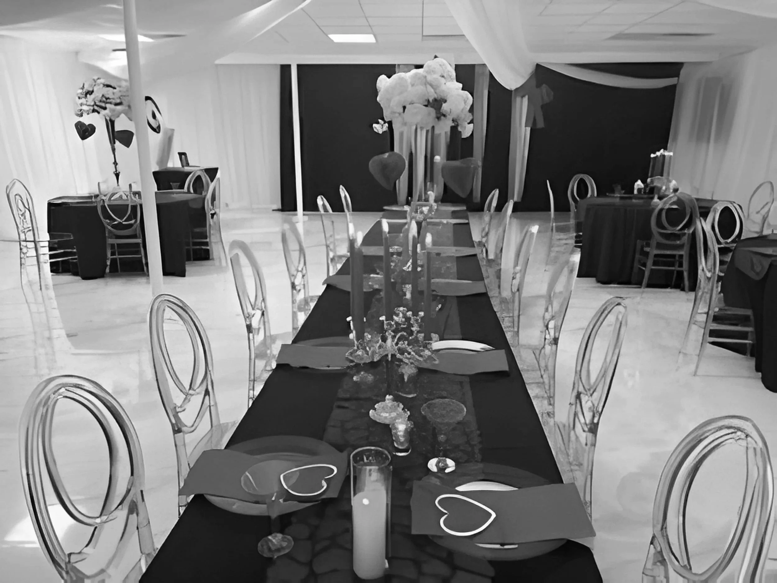 Black and white image of a long banquet table styled for a social event with  linens, table runners, candles, and clear acrylic chairs in a decorated event space