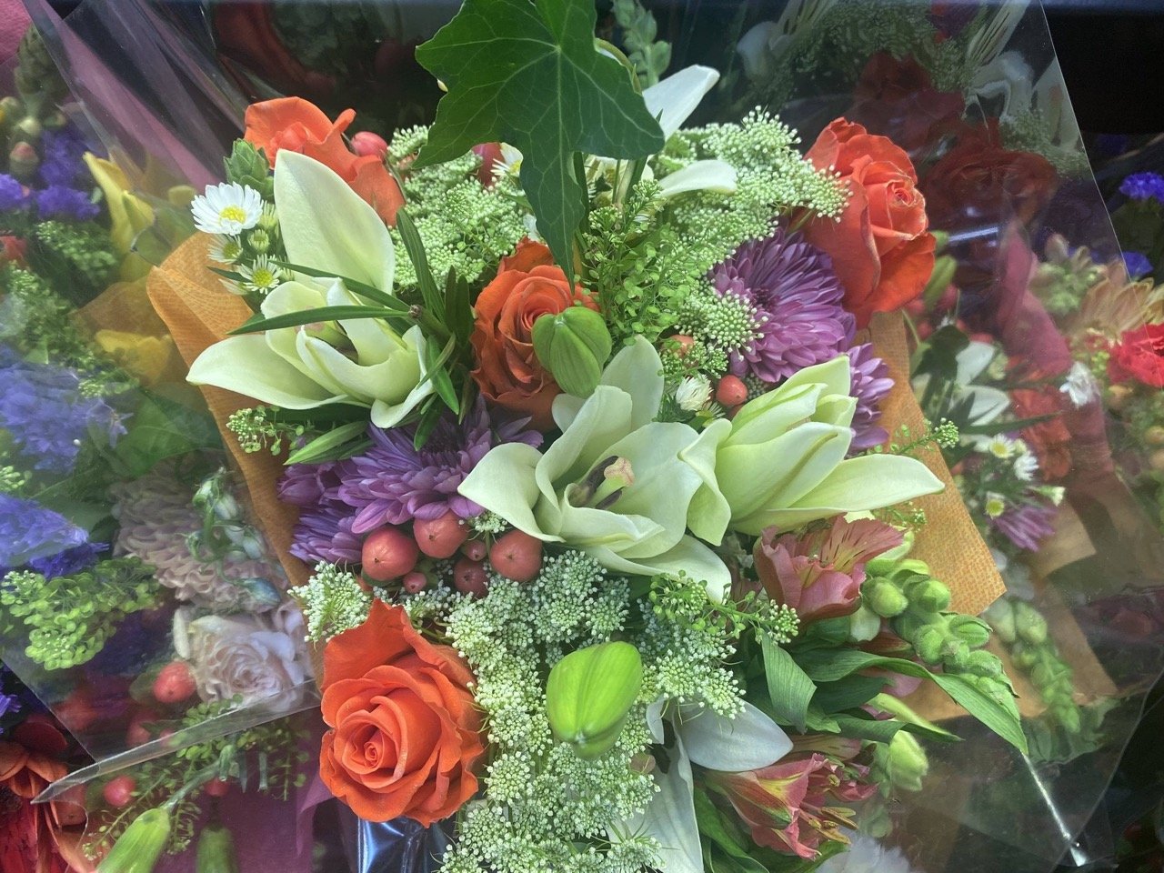 Hand-tied bouquet featuring orange roses, white lilies, purple mums, and greenery wrapped for a social event