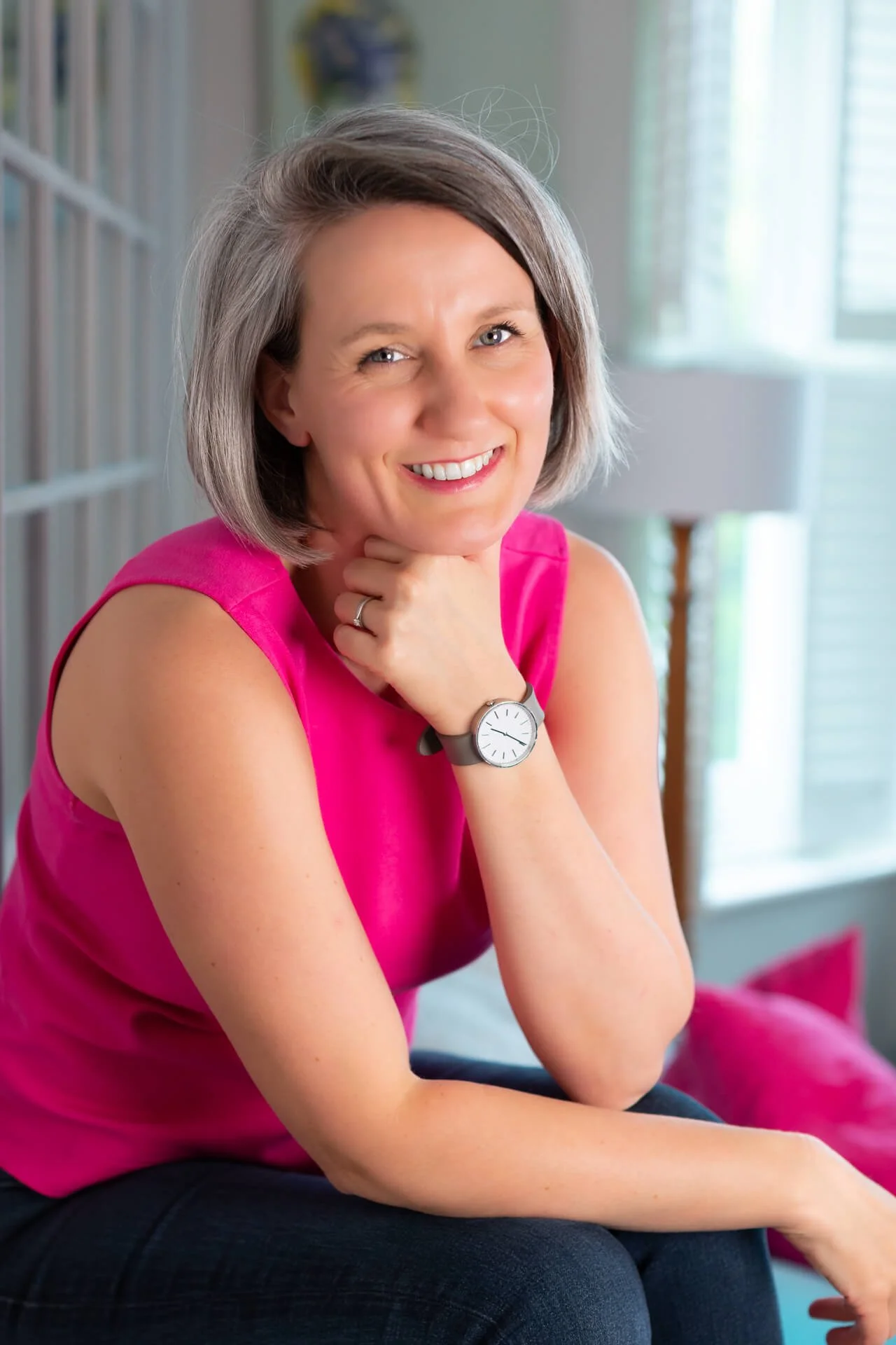 Jo Lofthouse sits in front of a mirror, in a pink top, with her chin on her hand, smiling