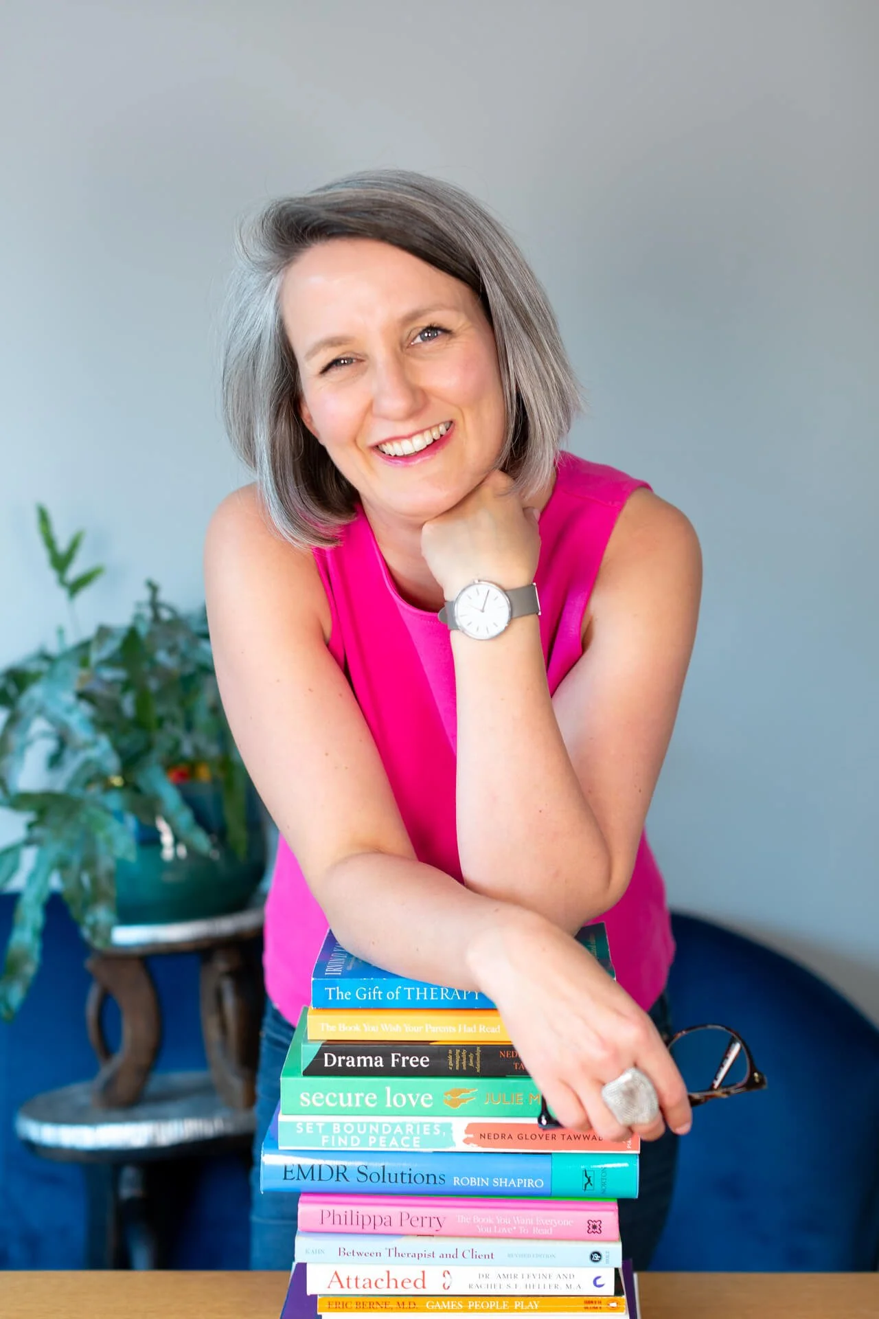 Jo leans on a pile of therapy books holding her glasses in one hand, wearing a pink top. She is smiling.