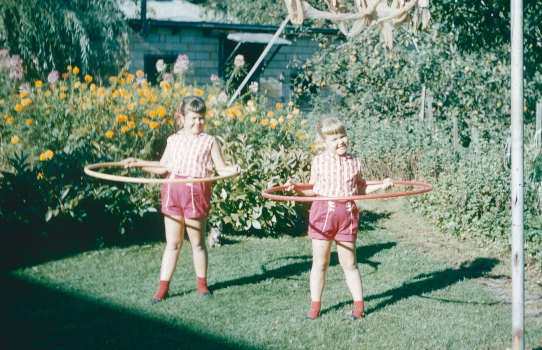 The image shows two young girls smiling and playing with hula hoops in a sunny, green garden during the mid-20th-century toy craze