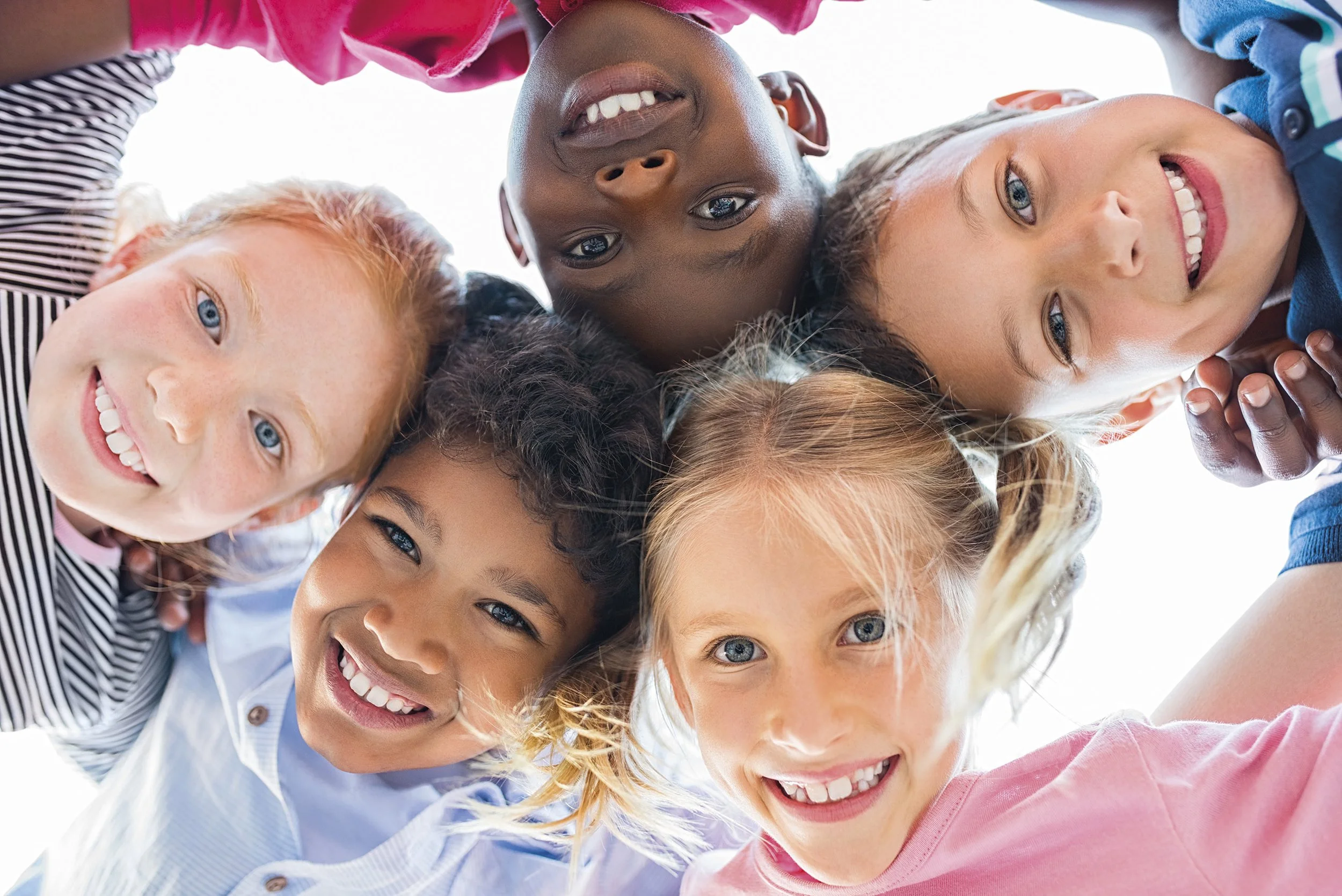 A photograph of 5 young children of mixed gender and race smiling in a huddle
