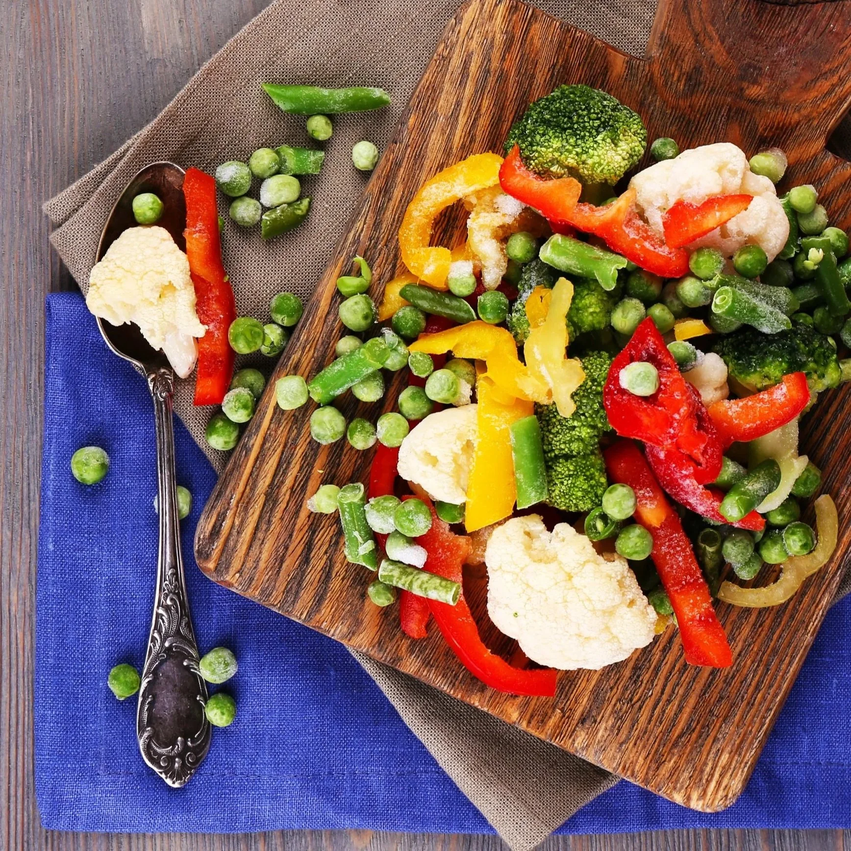 A wooden board with a colorful assortment of steamed vegetables including cauliflower, broccoli, red and yellow bell peppers, green beans, and green peas. A silver spoon with extra vegetables rests beside the board on a blue cloth, with additional peas scattered around.