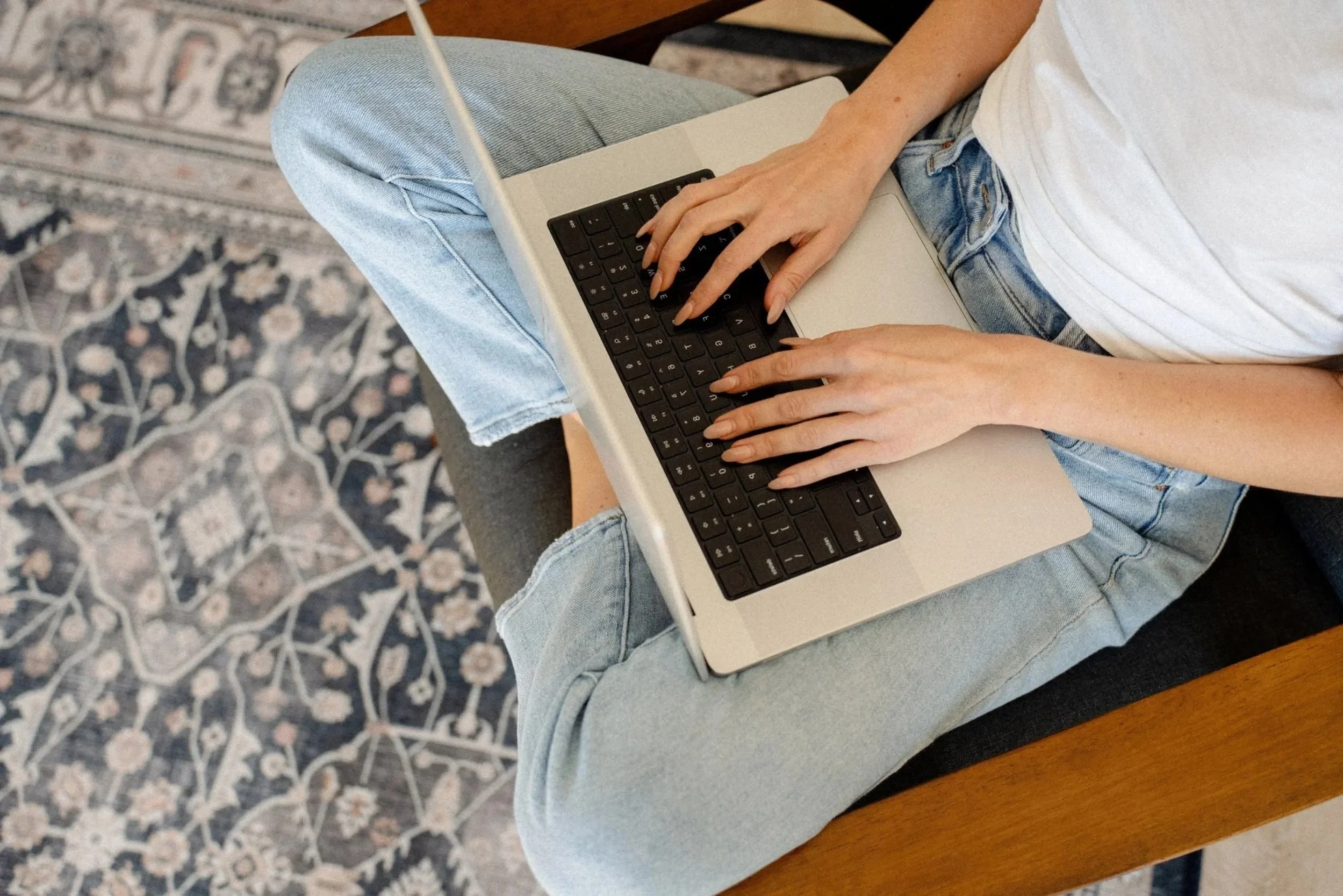 Person sitting on a wooden bench, wearing light blue jeans and a white t-shirt, using a silver laptop with black keys.
