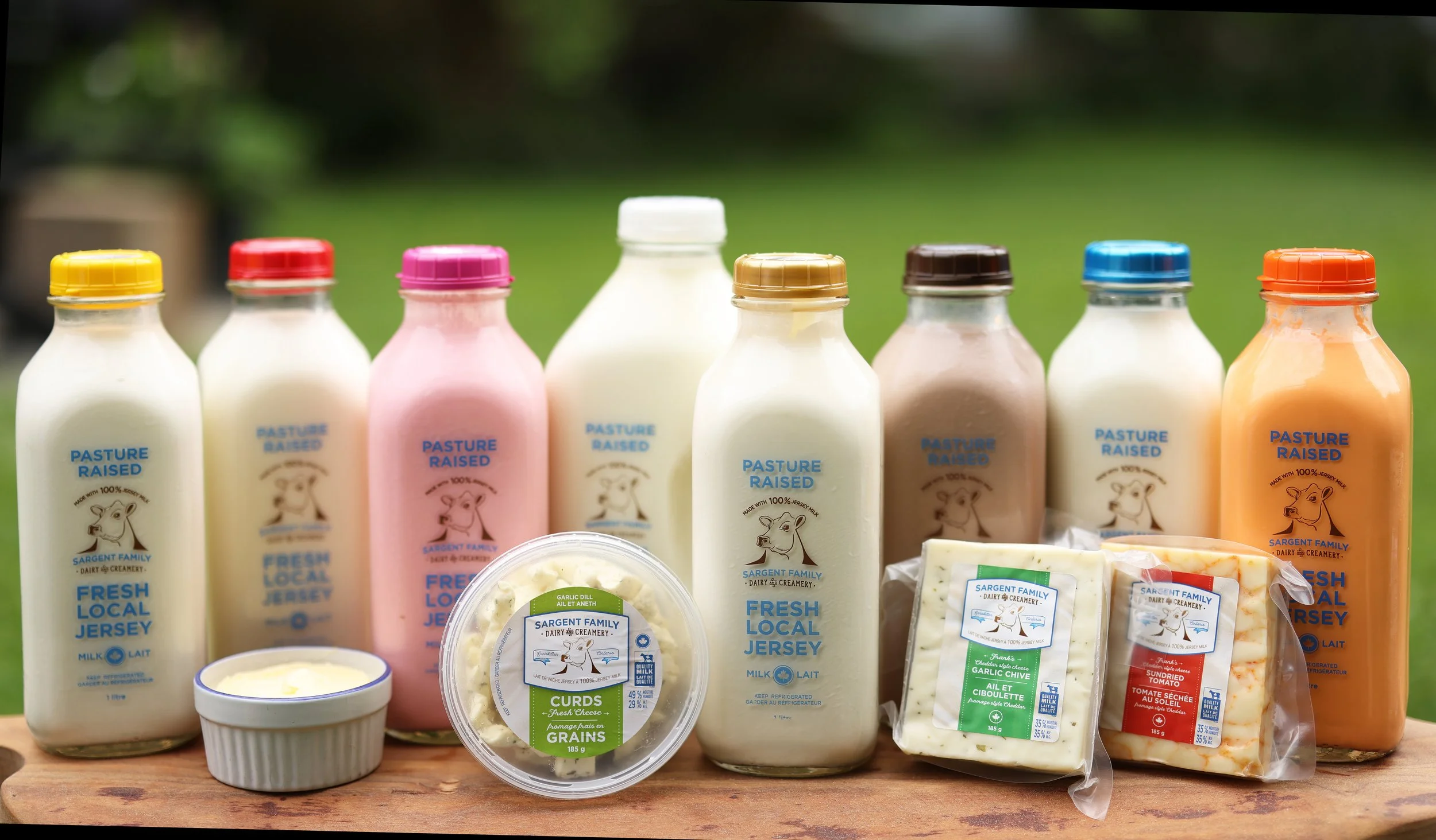 Six bottles of milk, two blocks of cheese, a small bowl of butter, and two packages of cheese slices arranged on a wooden surface outdoors.