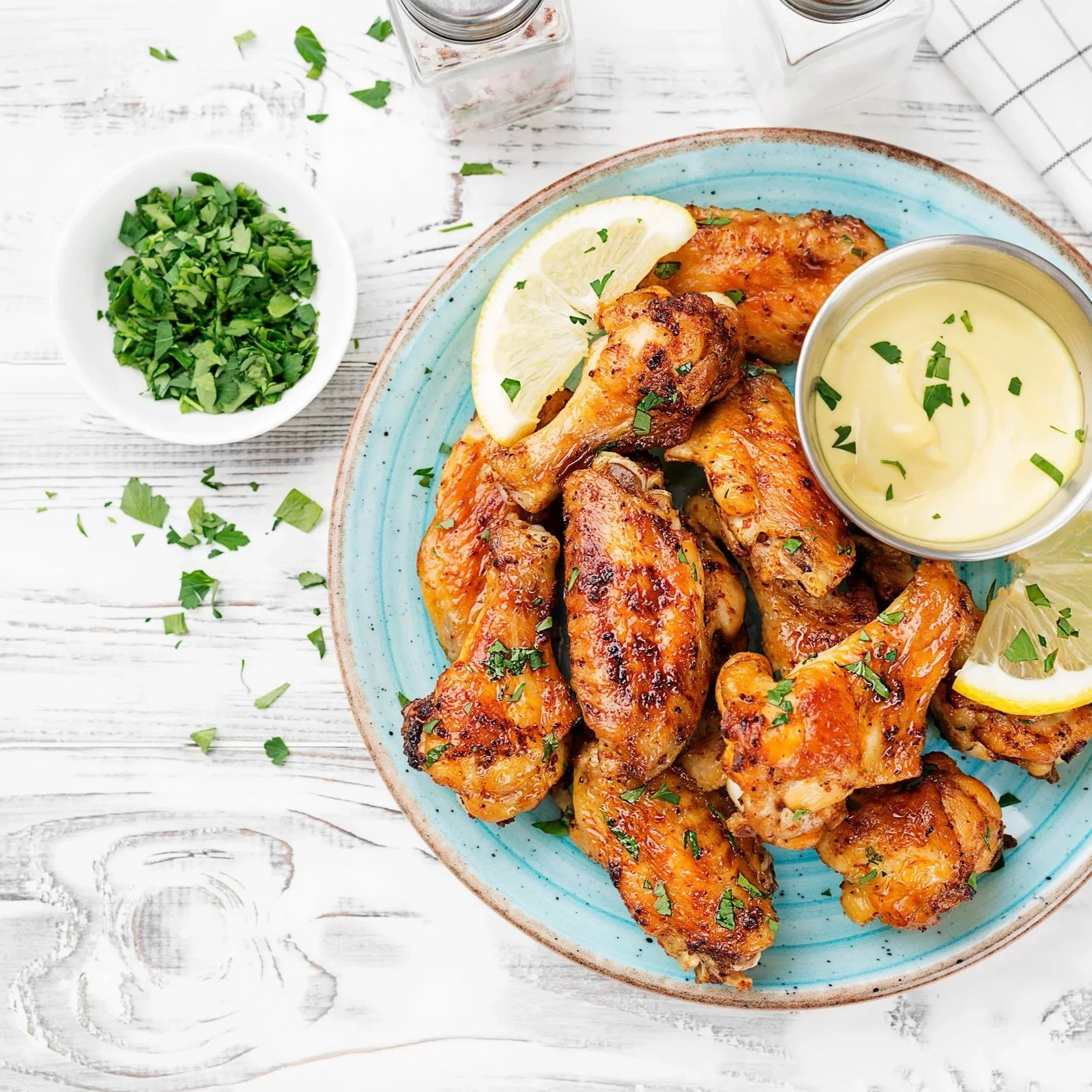 Plate of cooked chicken wings garnished with chopped parsley and lemon wedges, with a small bowl of creamy dipping sauce and fresh chopped herbs on the side.