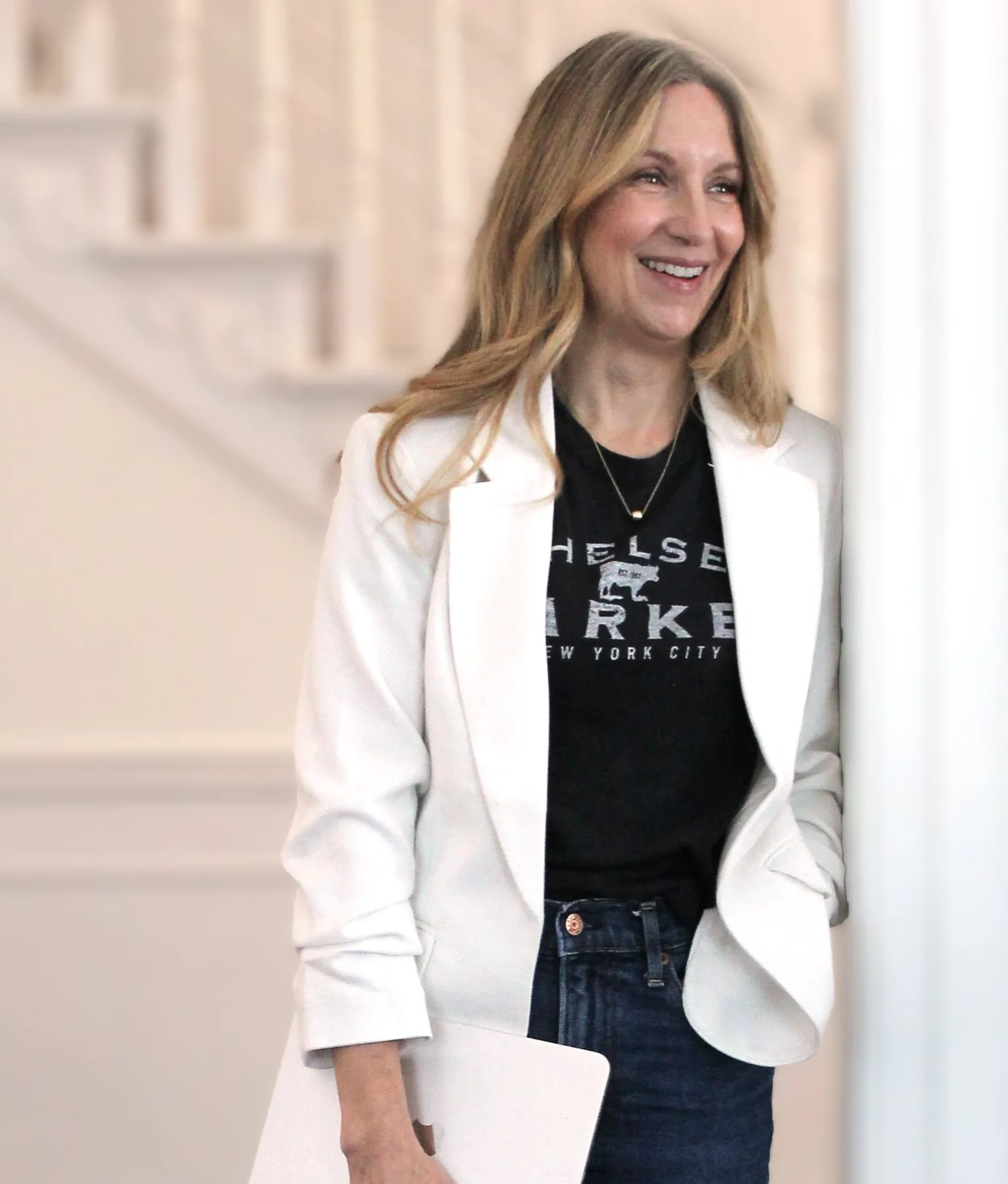 Woman with long blonde hair smiling, wearing a white blazer over a black t-shirt with white text, dark jeans, and holding a white laptop, standing indoors.