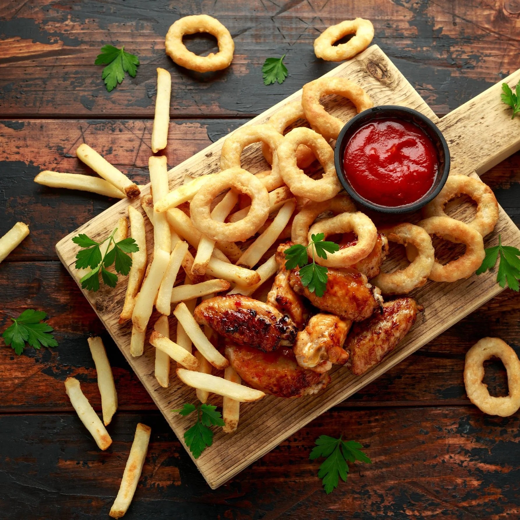 A wooden serving board with fried onion rings, French fries, grilled chicken wings, plenty of parsley, and a small bowl of ketchup on a rustic wooden table.