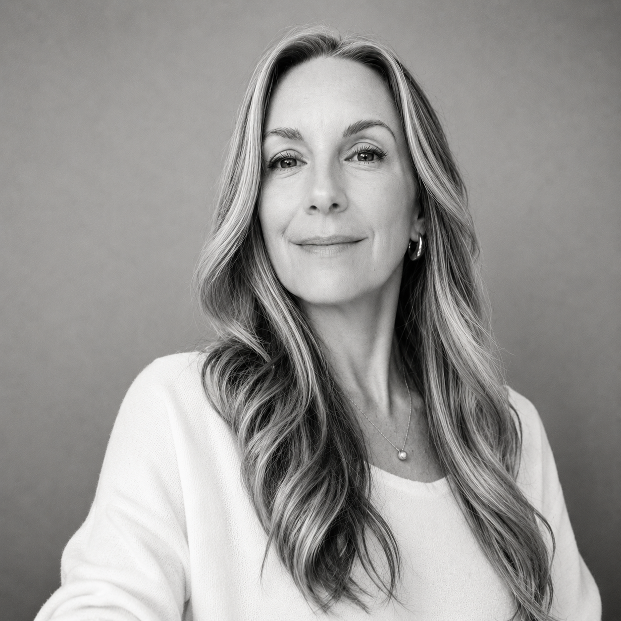 Black and white portrait of a woman with long wavy hair, smiling softly, wearing a light-colored top, earrings, and a necklace, against a plain gray background.