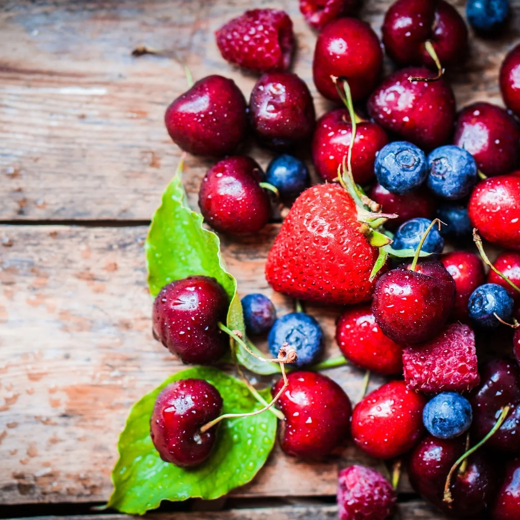 A variety of fresh berries including cherries, blueberries, and a strawberry on a wooden surface with a green leaf.