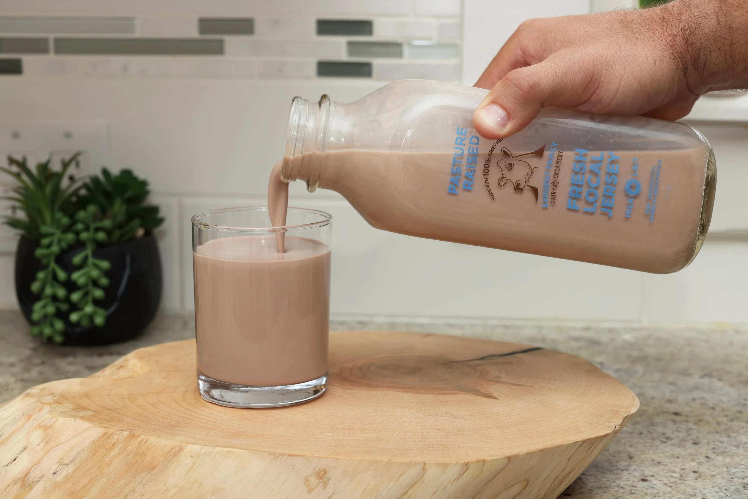 Person pouring chocolate milk from a glass bottle into a glass on a wooden surface in a kitchen.