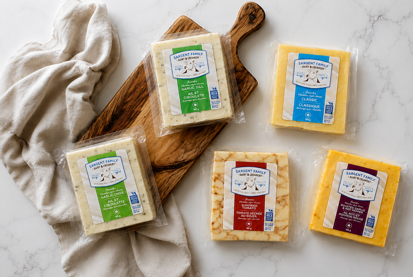 Five blocks of assorted cheese on a white marble surface, including garlic dill, garlic chive, sun-dried tomato, classic, and roasted garlic and red pepper varieties, with a beige cloth and a wooden cutting board.