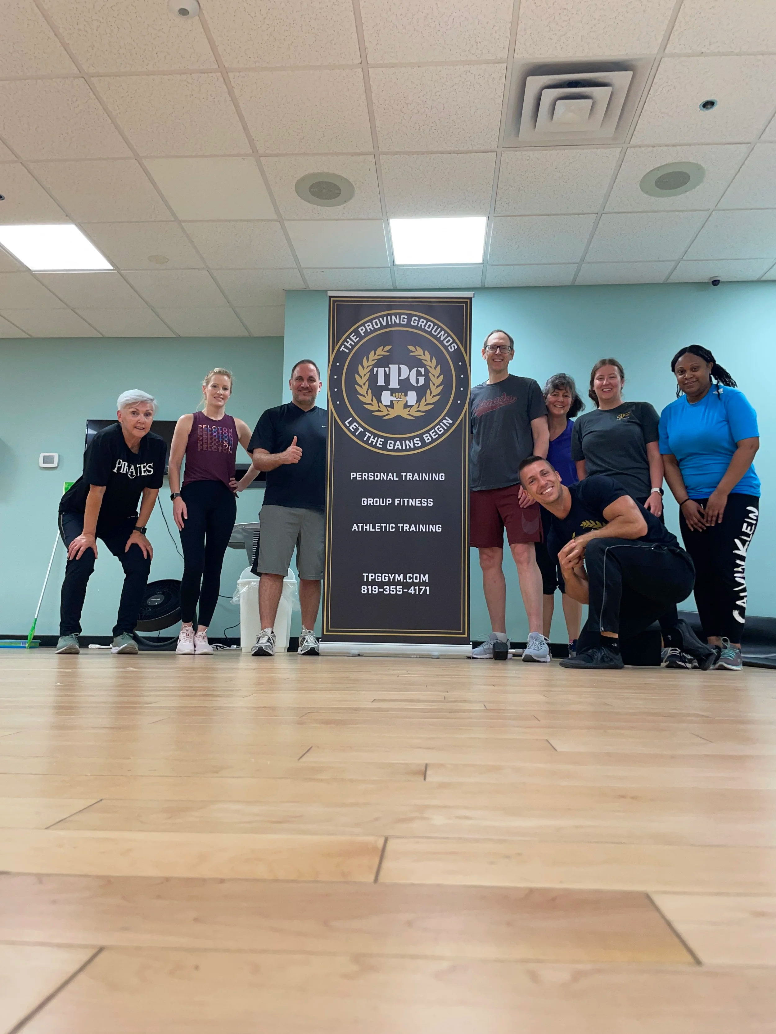 Group of eight people standing in fitness studio behind a large vertical banner for TPG Gym, with some smiling and posing casually.
