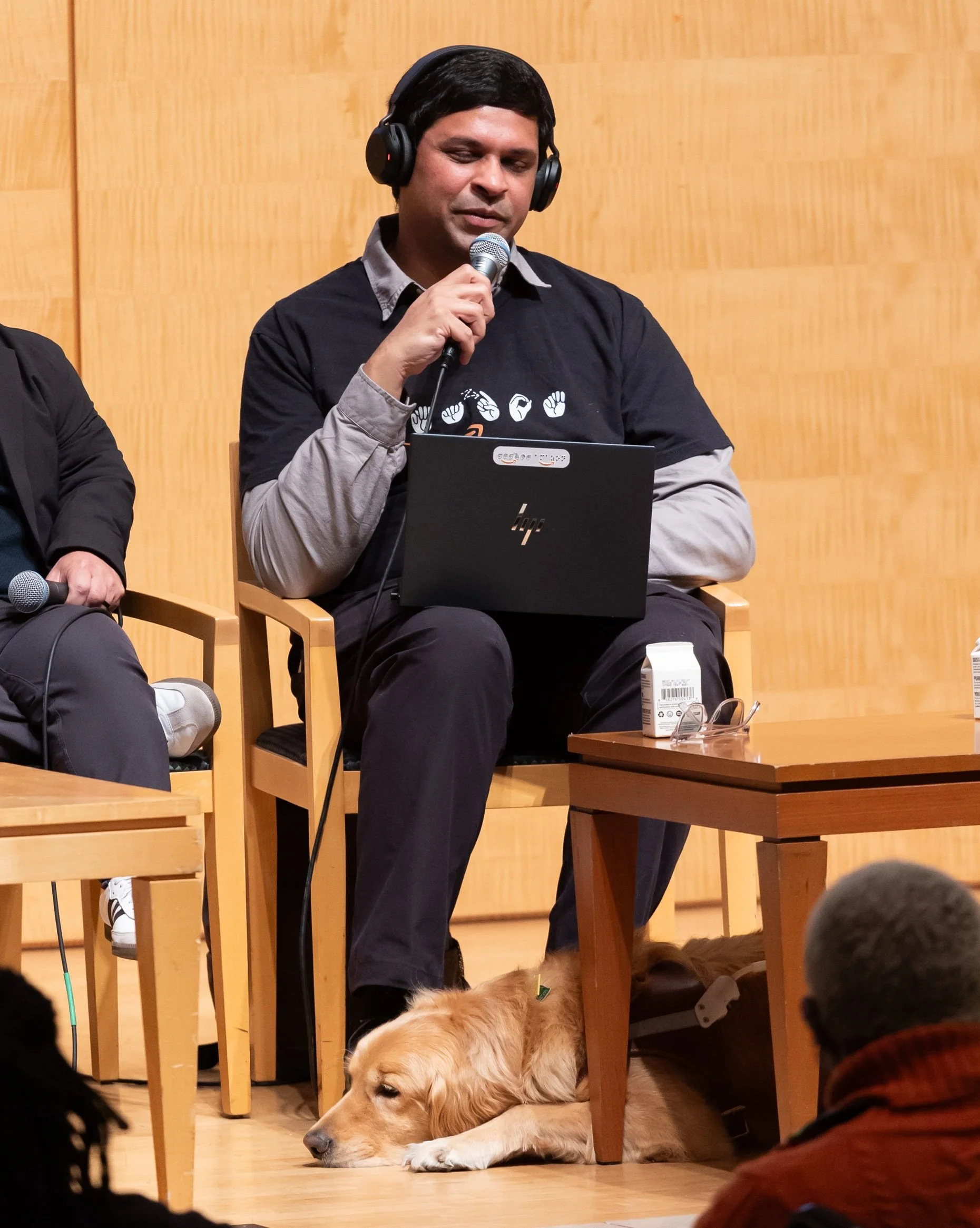 Blind man sitting with a laptop on his lap, waering a headset and speaking in a microphone while his service golden retriever lays down at his feet