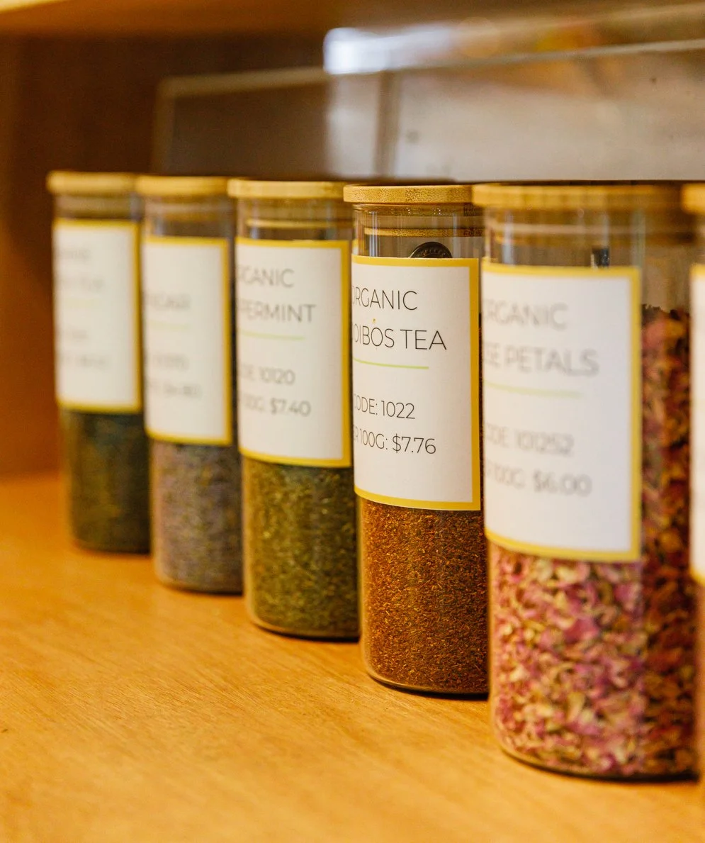 A row of clear glass jars with wooden lids containing different dried herbs and teas, labeled with white tags showing their names and prices. The jar in the center is labeled "Organic Hibiscus Tea."