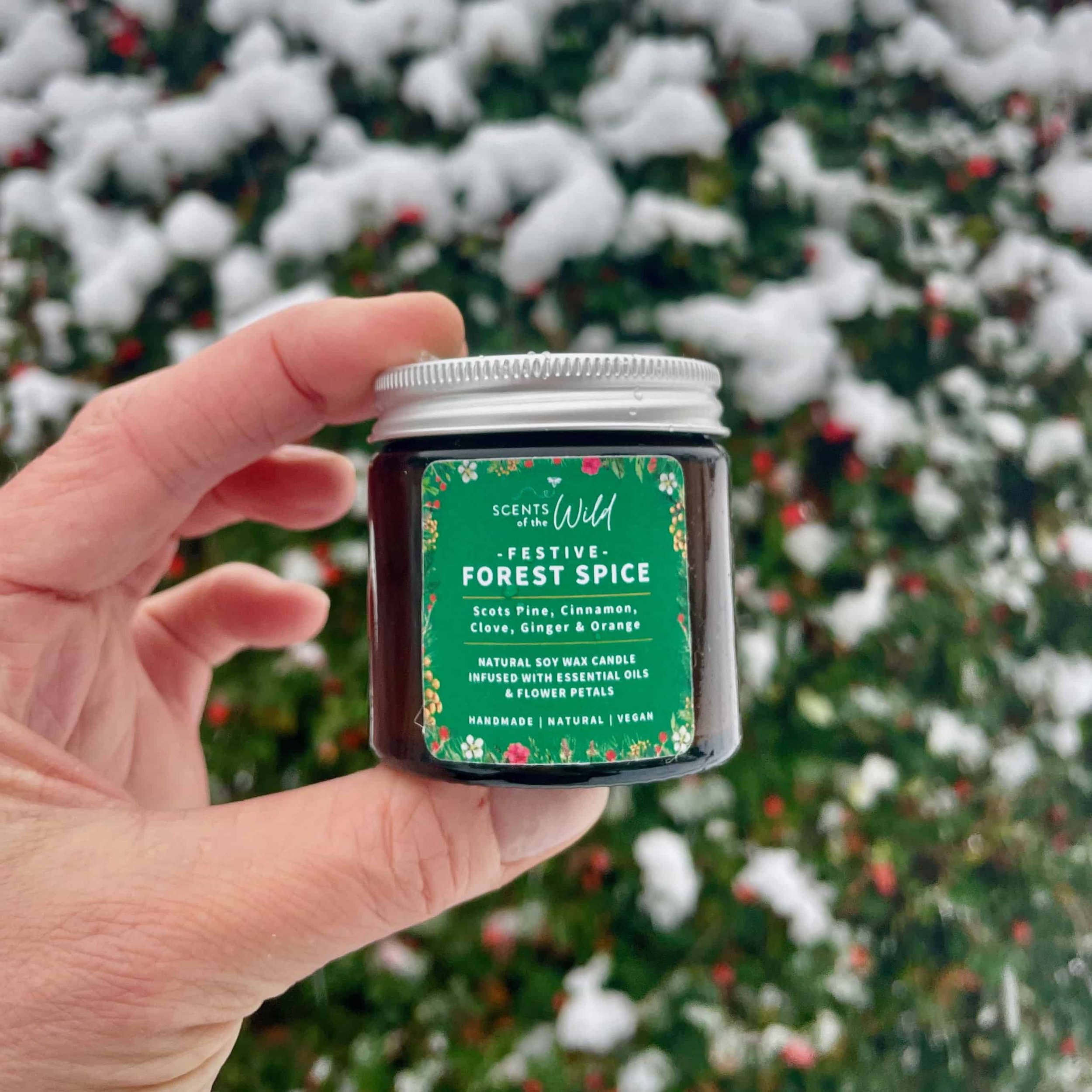 Small festive candle jar held in front of snowy bushes.jpg