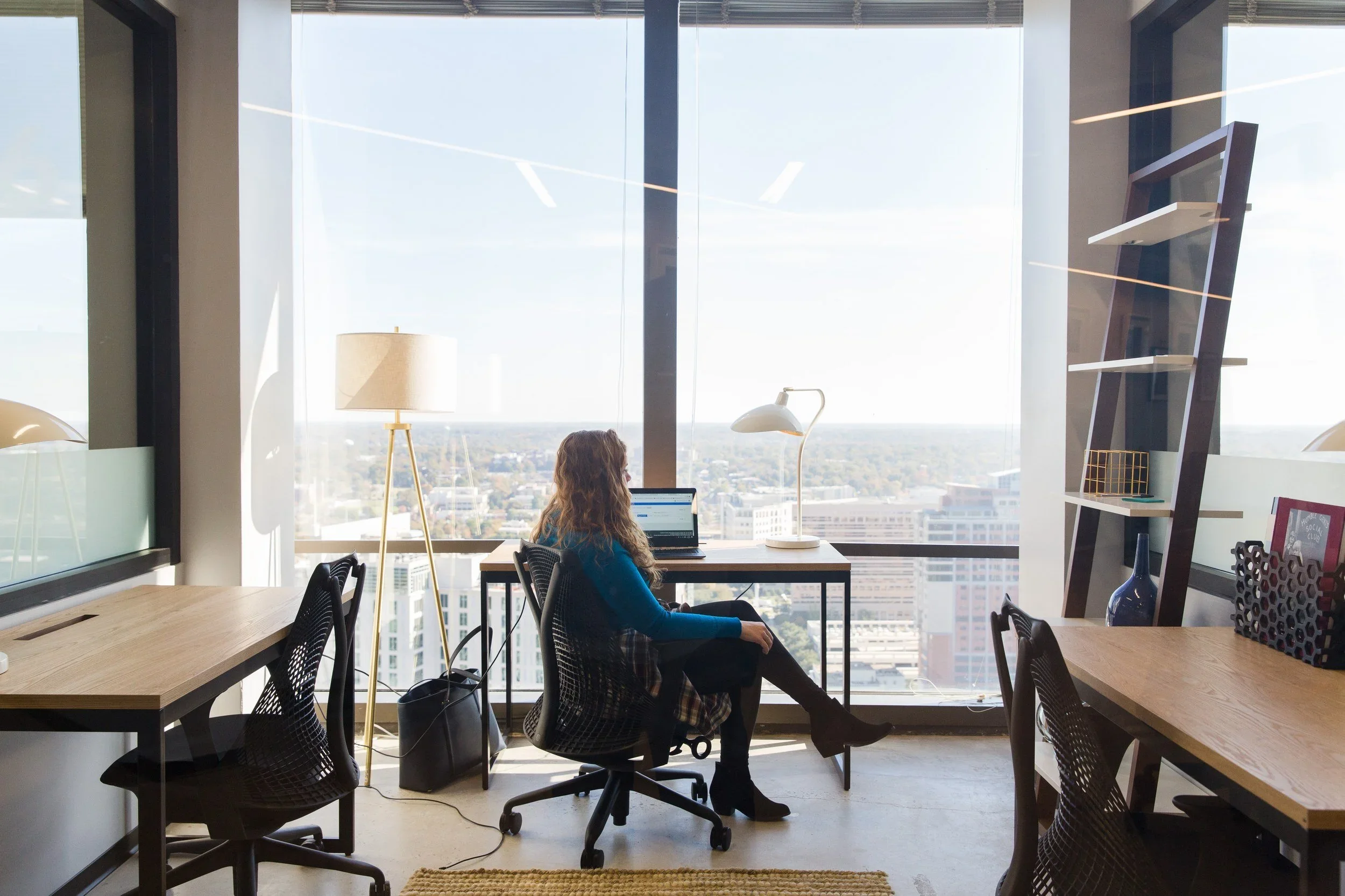 Woman working on a laptop at a desk by a large window