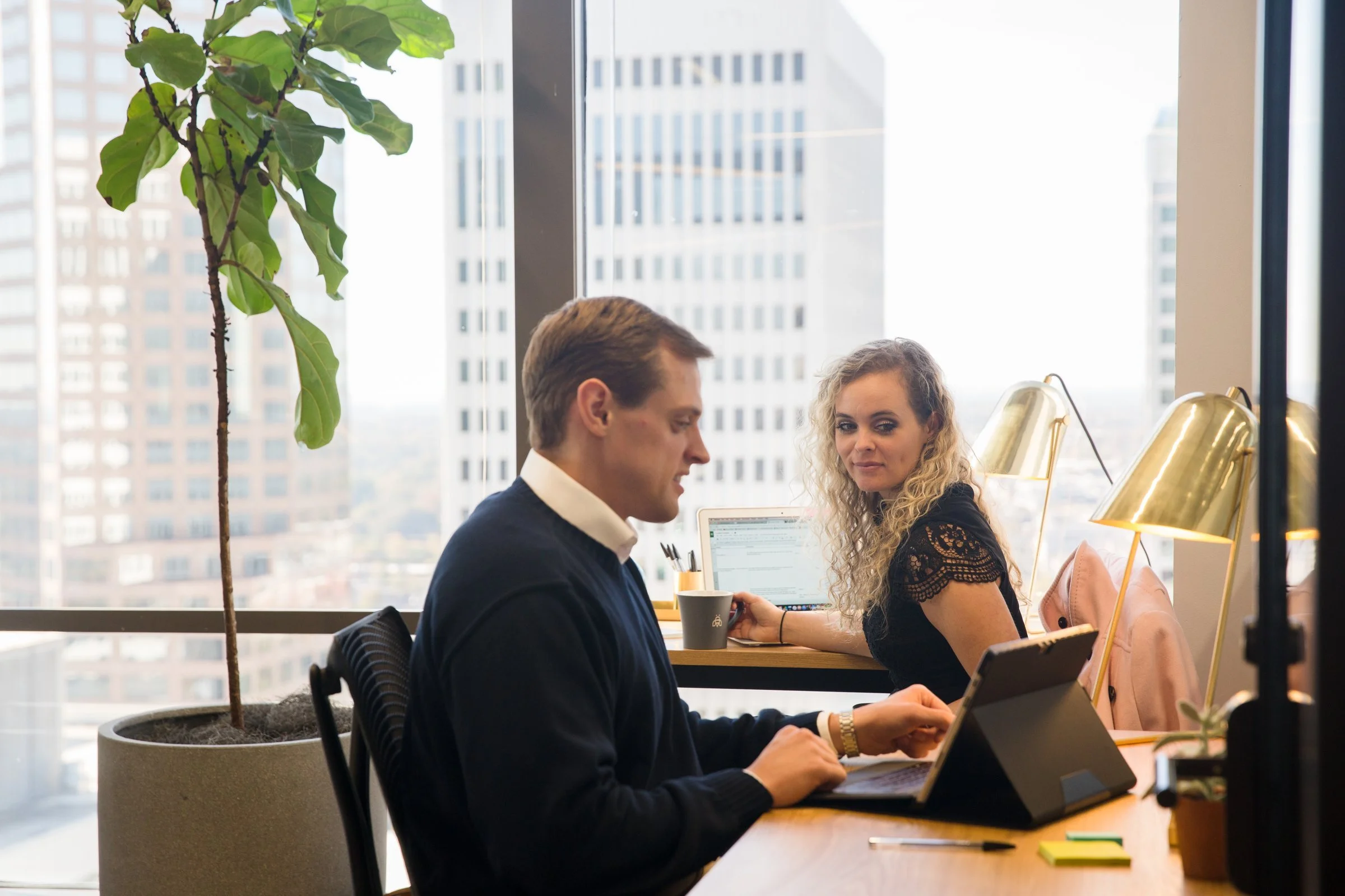 Two colleagues at a Charlotte office window desk working on an iPad and laptop, Apple devices managed through Apple Business Essentials