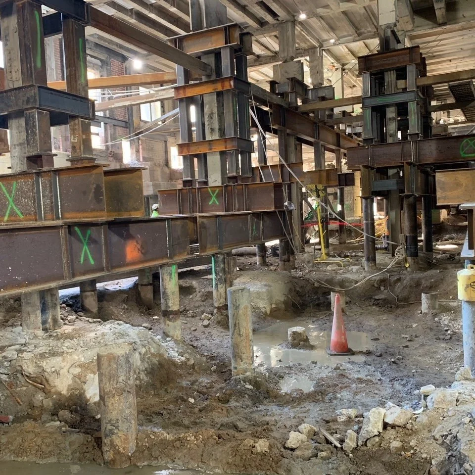 Construction site with steel beams, scaffolding, and concrete piles in an unfinished building interior.