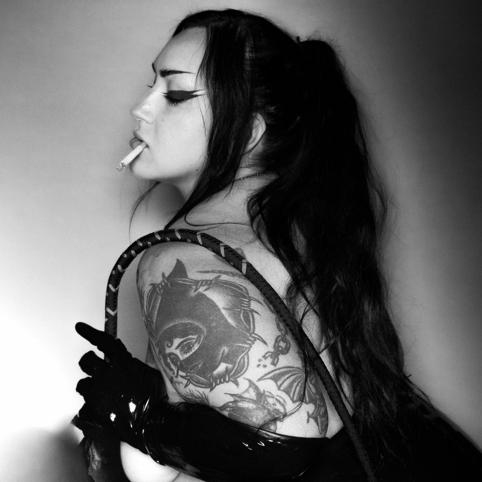 Black and white photo of a woman with long dark hair, holding a cigarette in her mouth, wearing a tattoo sleeve, glossy gloves, and a strap over her shoulder.