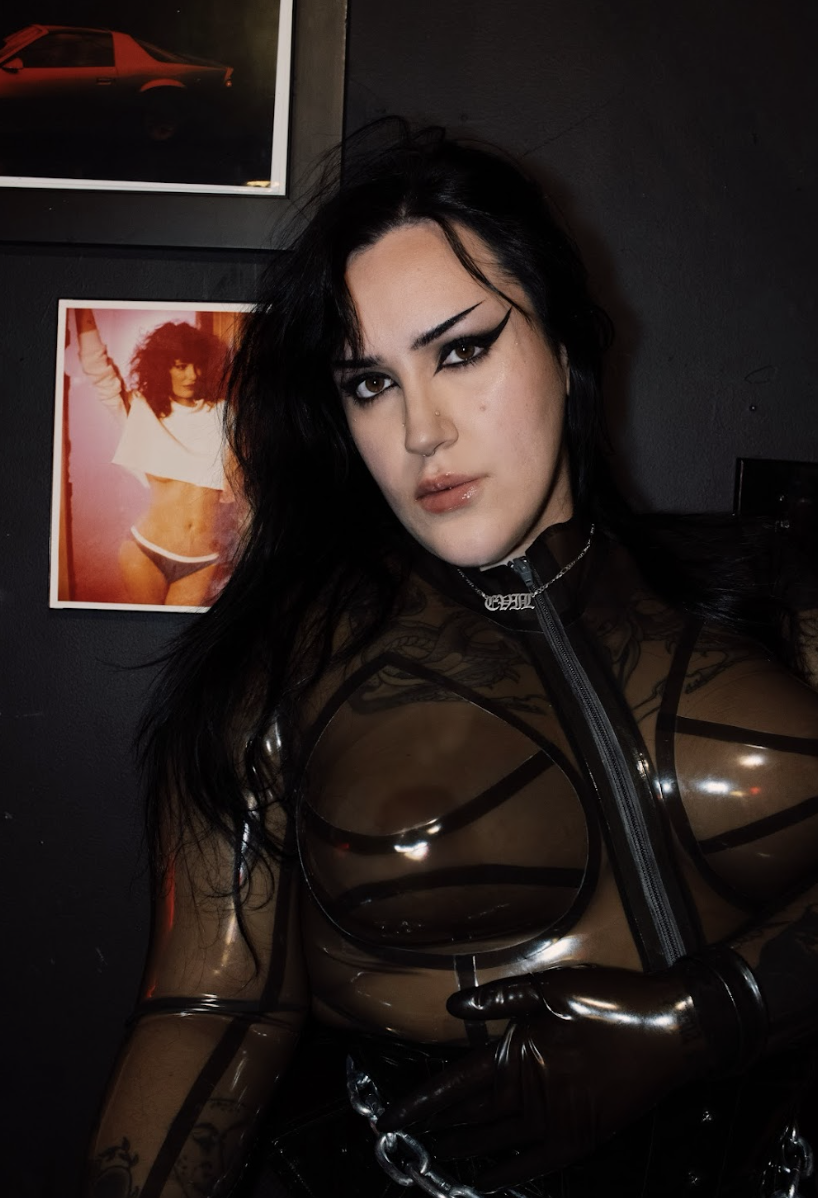 A woman with black hair and dramatic makeup wearing a see-through black latex top and gloves, standing indoors with framed posters on the dark wall behind her.