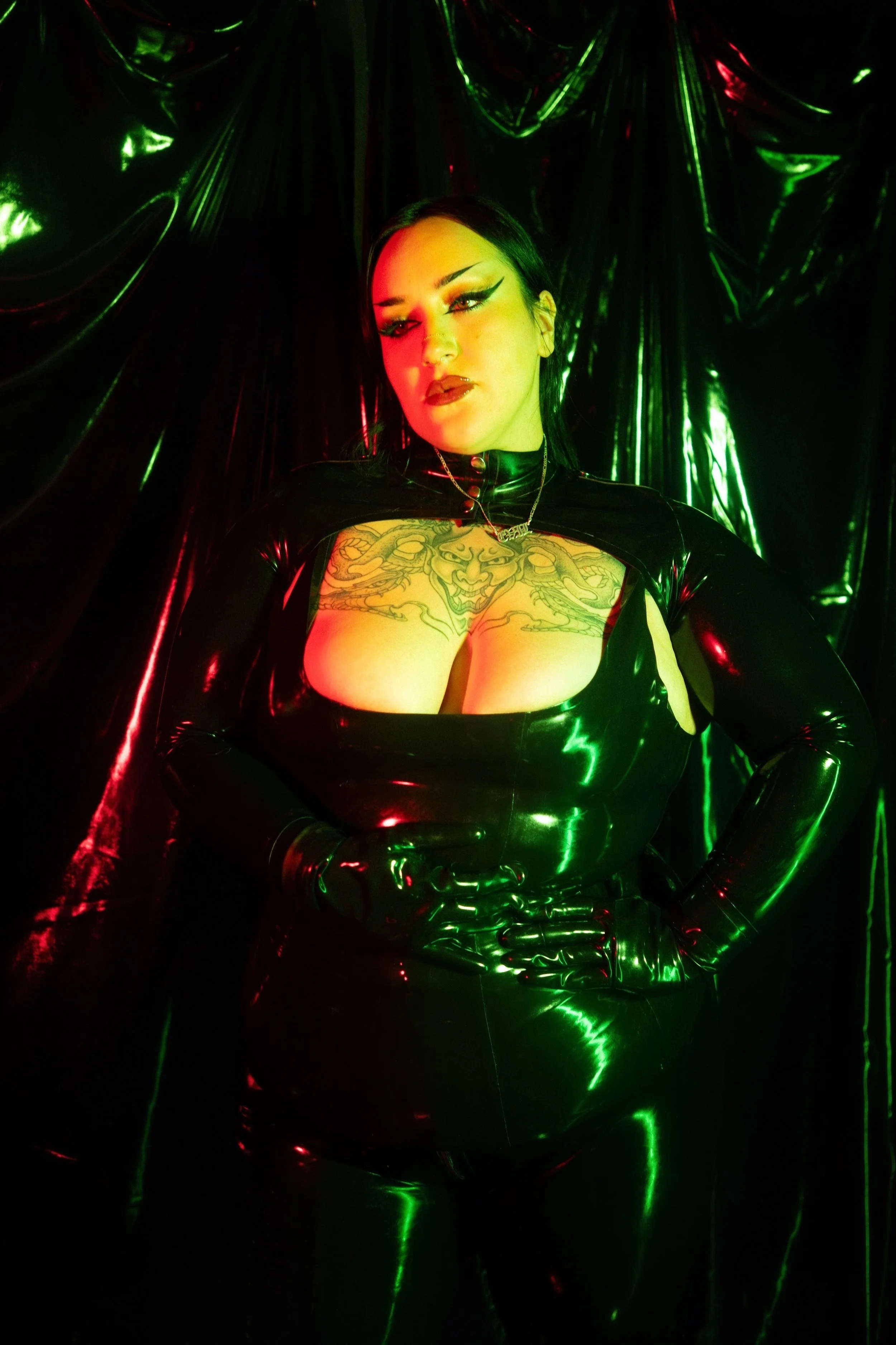 Mistress Micha Malvada dressed in a latex catsuit, latex gloves, and a latex bolero. She is shown in front of a shiny black background. She poses With her hands across her waist while her eyes gaze into the distance. The scene is lit in green lights.