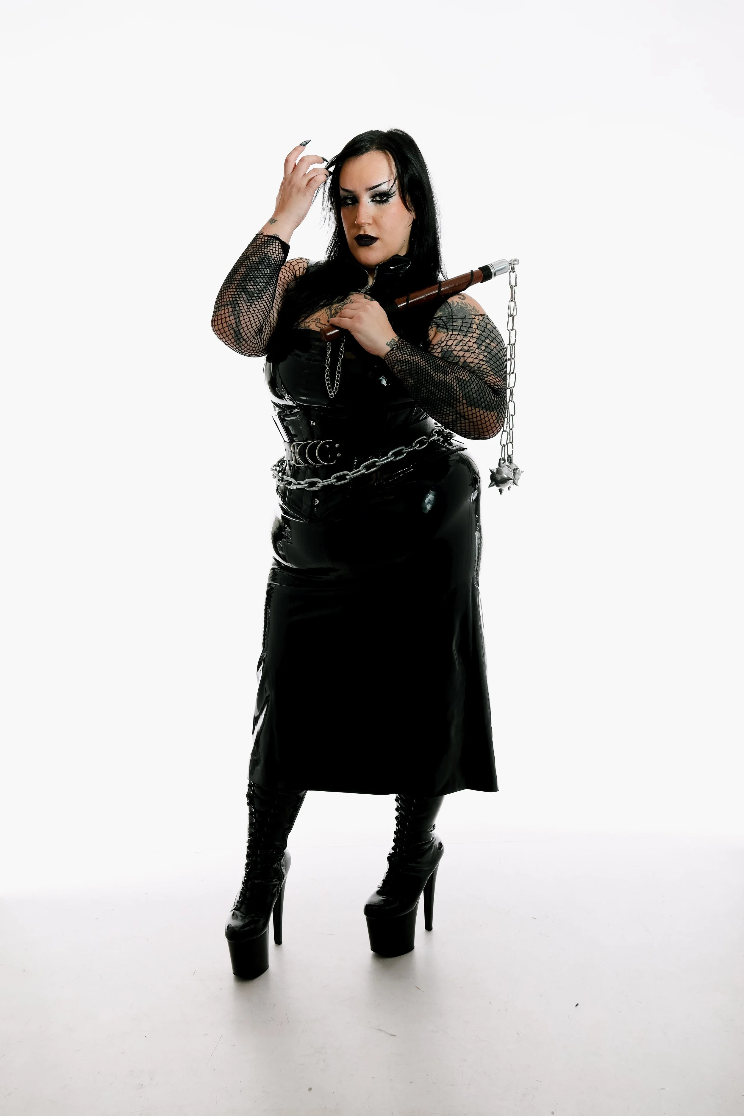 A woman dressed in a black latex dress with fishnet sleeves, wearing high platform boots, holding a spiked flail, standing against a plain white background.