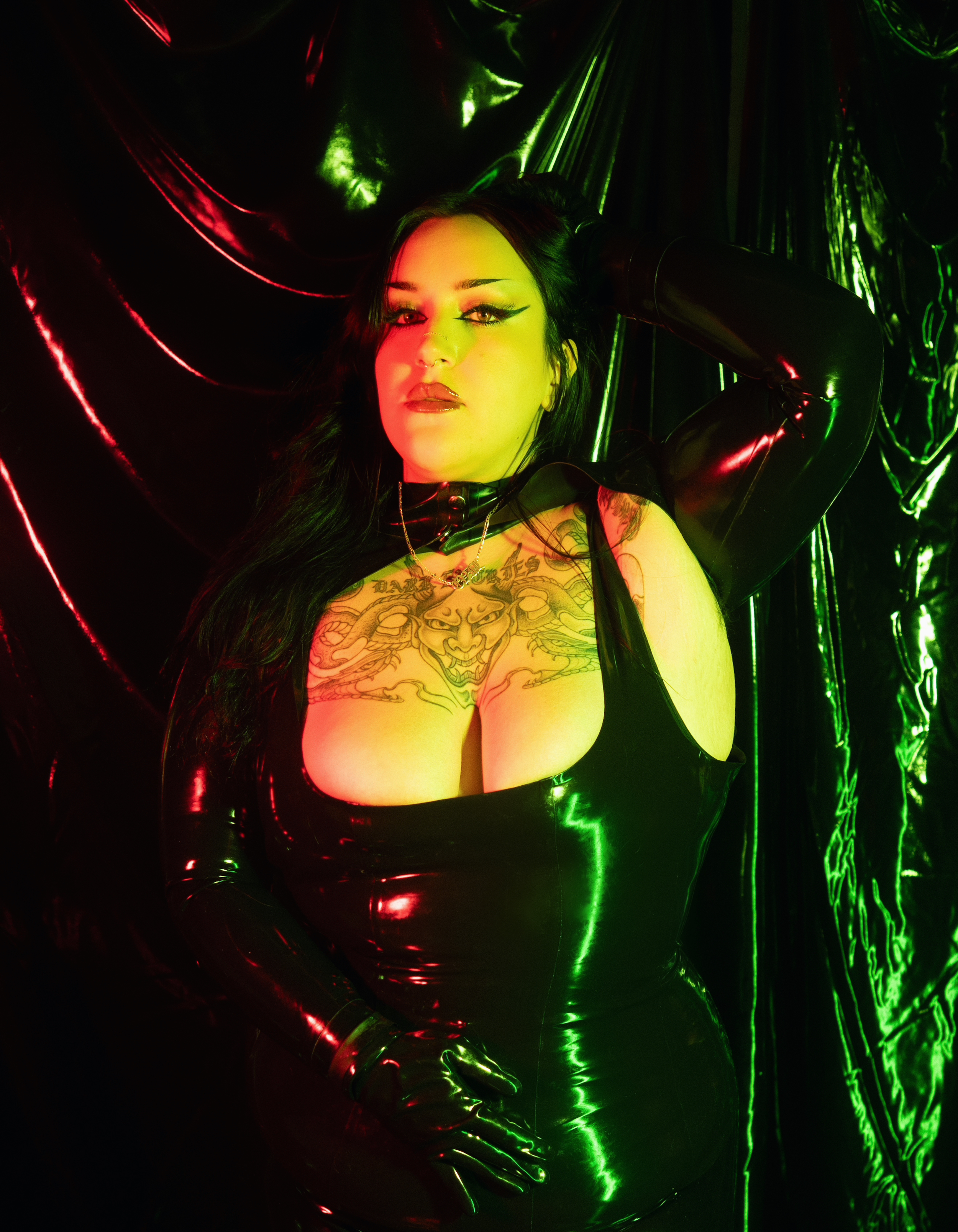 A woman with long dark hair, tattoos on her chest, wearing glossy black latex clothing and gloves, posing in front of a shiny black and green background.