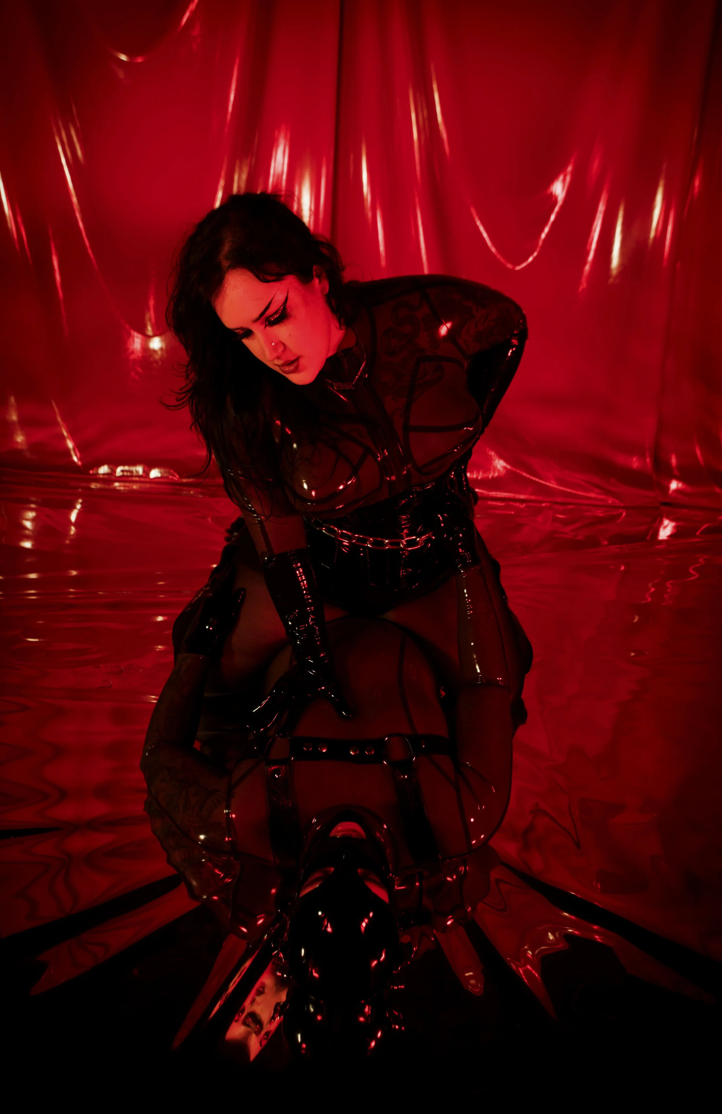 A woman dressed in black latex and fishnet clothing, kneeling on a shiny, reflective floor in front of red, metallic curtains.