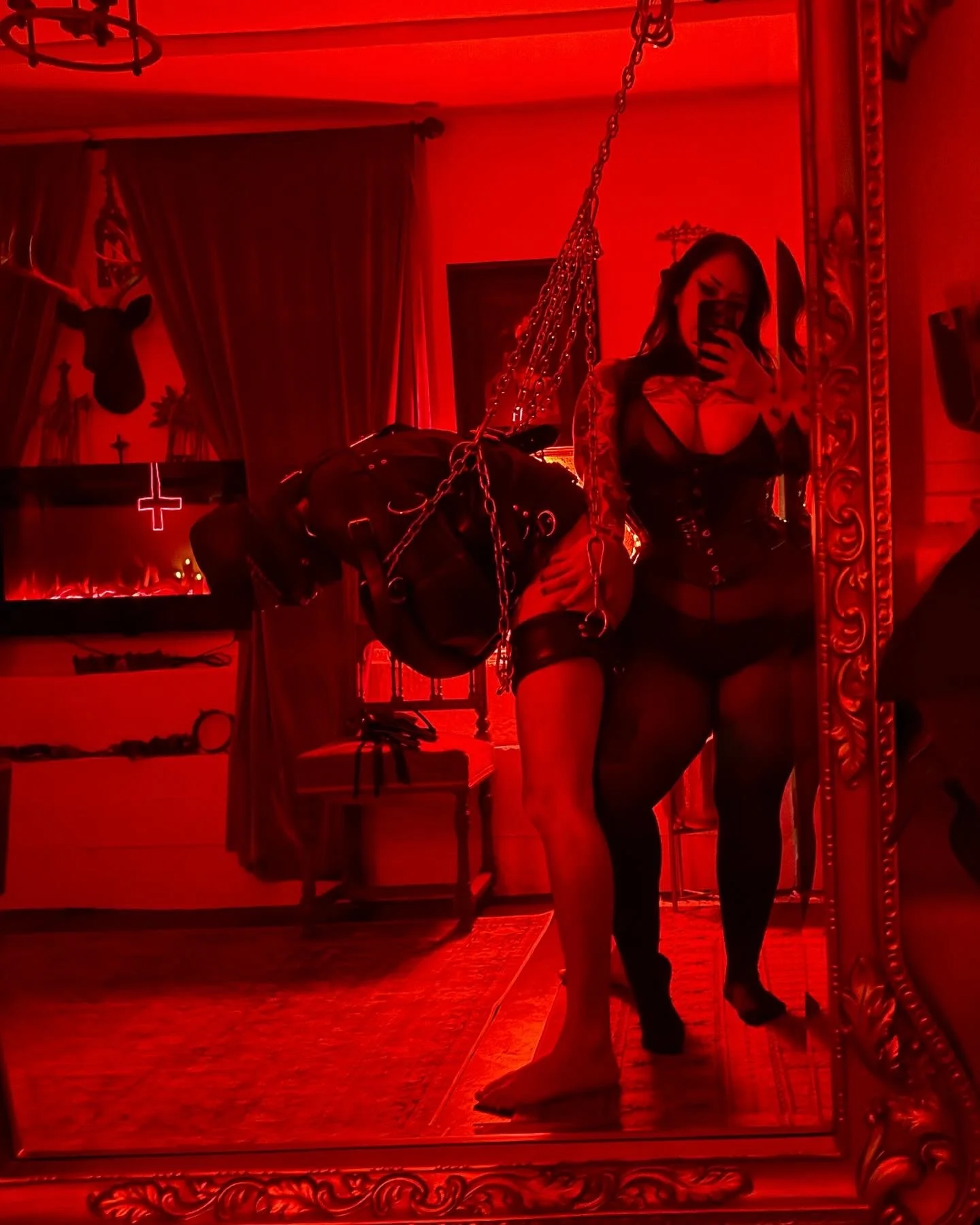 A woman taking a mirror selfie in a room bathed in red lighting. She is dressed in black lingerie and stockings, with tattoos visible on her arm. The room has decorations including a mounted deer head, a cross on the wall, and chain lighting.