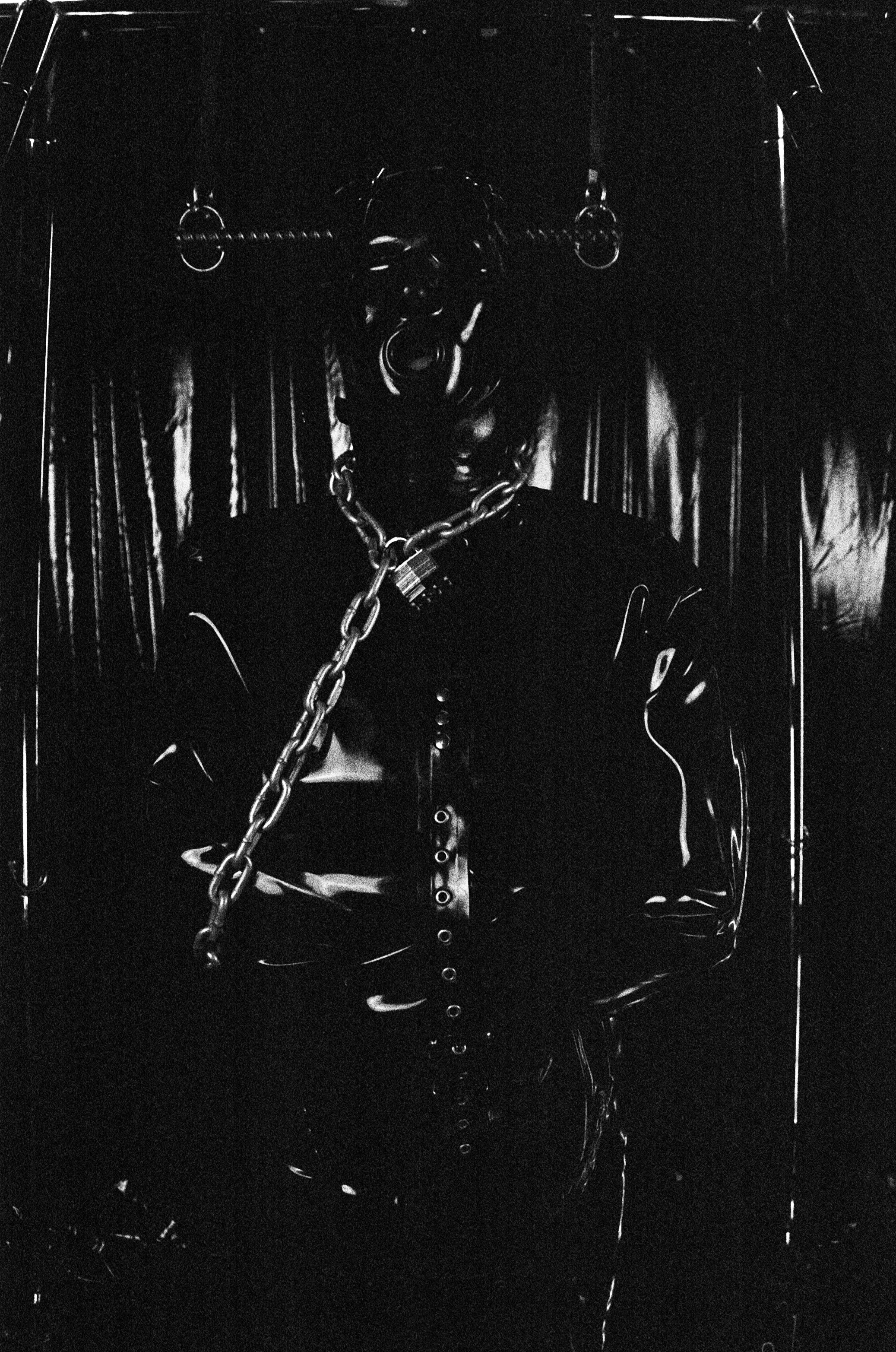 A figure wearing a shiny black outfit with metallic chains and jewelry, including necklaces and earrings, standing in front of a dark background with faint vertical lines.