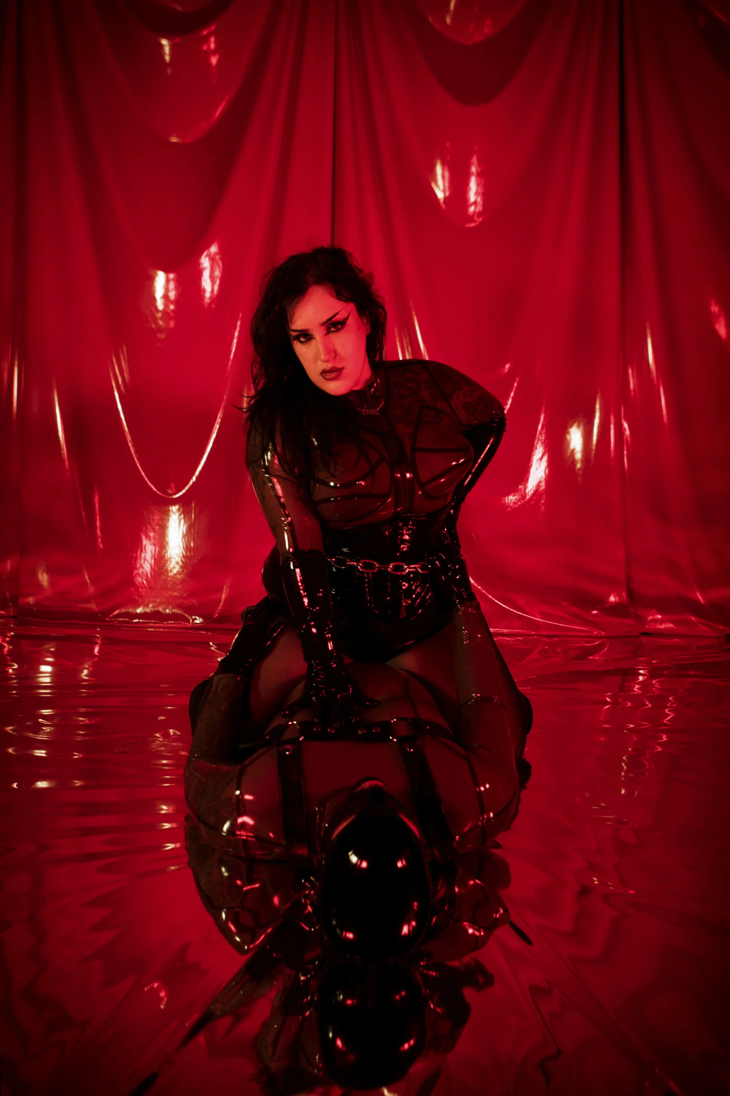 A woman with dark curly hair and makeup, wearing a black latex outfit with chains, kneeling on a reflective surface in front of a red satin backdrop, with a black reflective helmet or headgear on the ground.