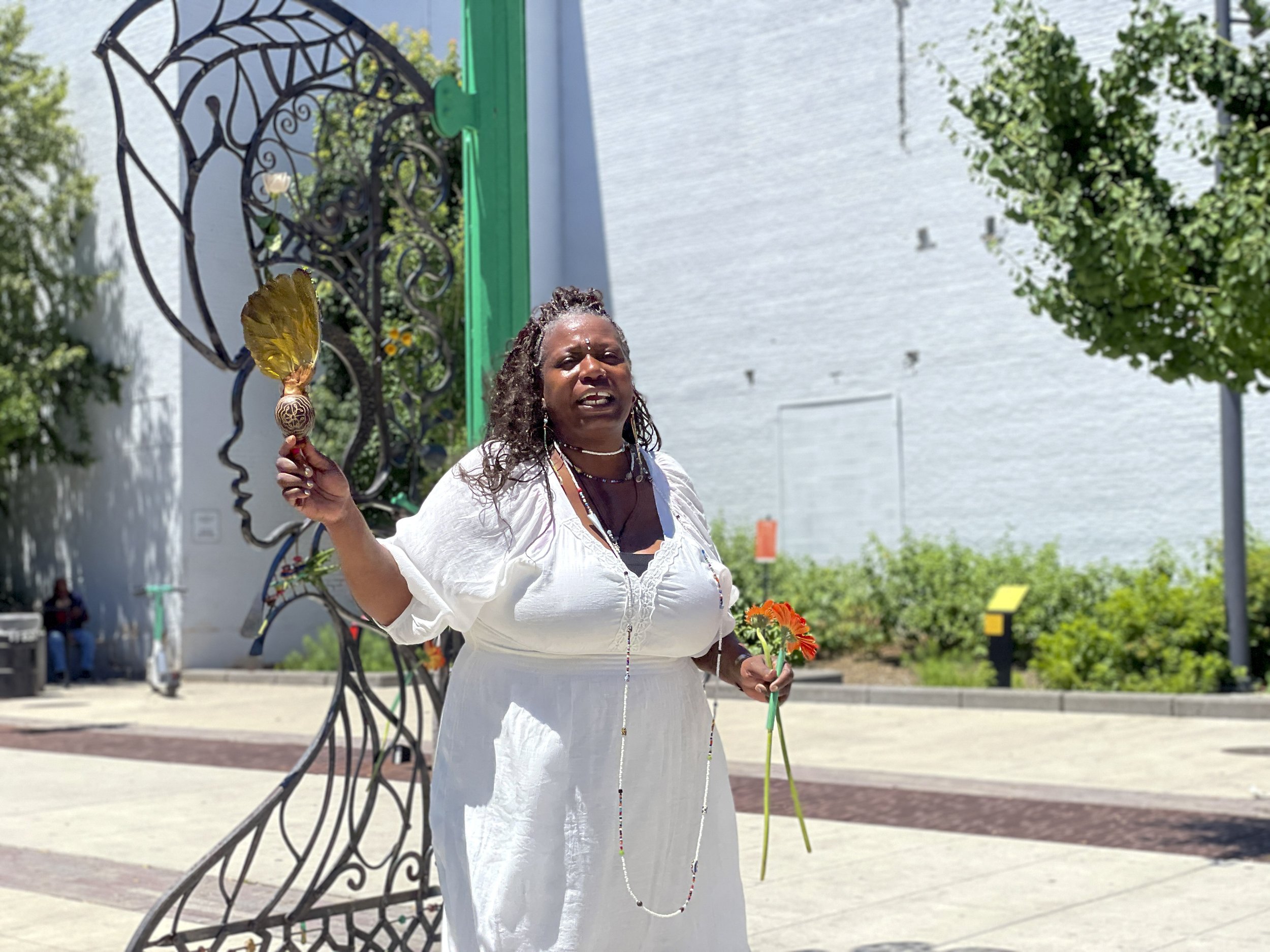 A black woman with long braids stands in front of a metal sculpture outdoors, singing.