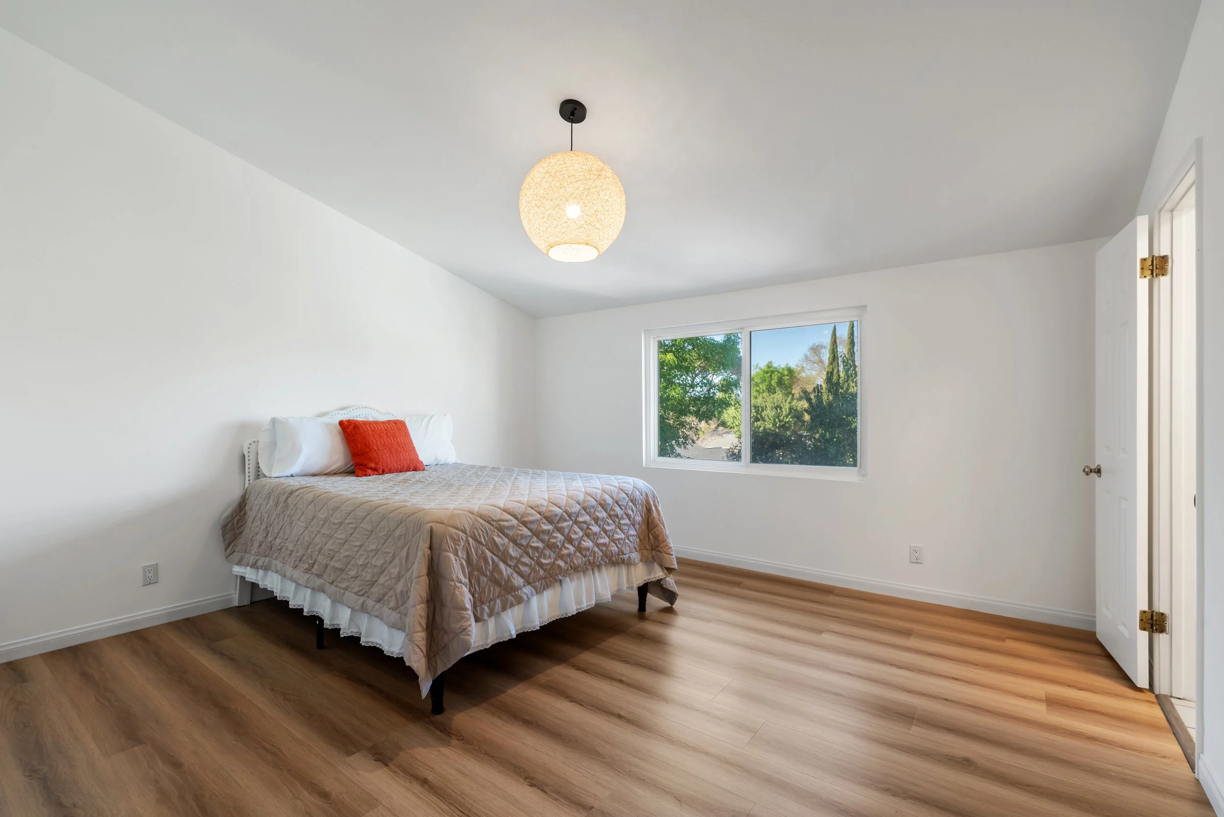 A bedroom with white walls, wood flooring, a bed with beige quilt, white pillows, and a red cushion. There's a window showing greenery outside and a ceiling light fixture.