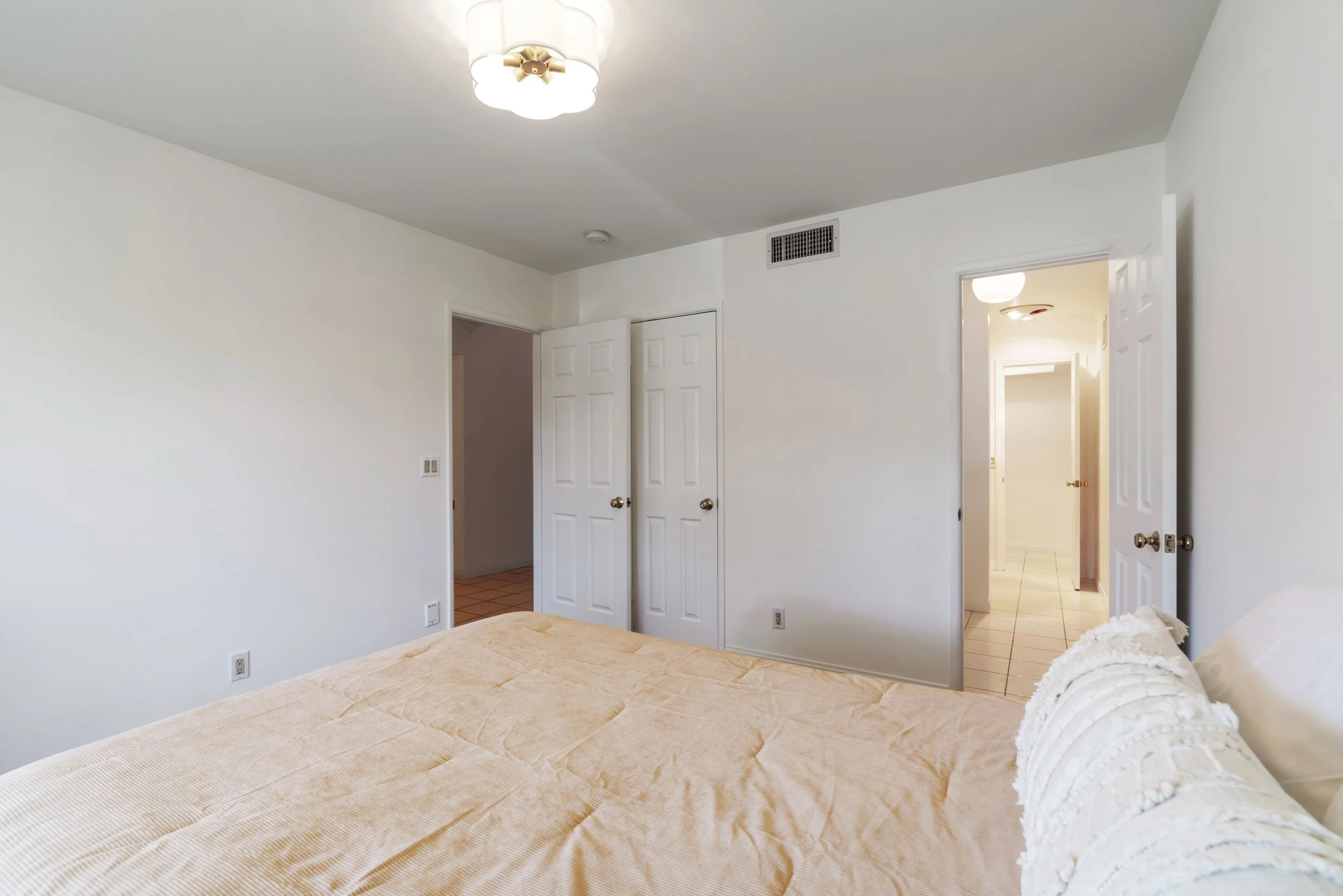 Empty bedroom with white walls, beige bedspread, and open closet doors, with doorway leading to a brightly lit hallway.