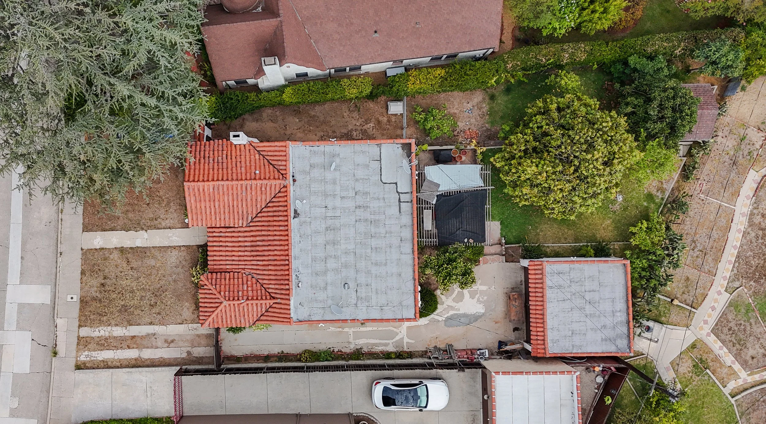 An aerial view of a residential neighborhood showing rooftops, a driveway with a parked white car, and a backyard with trees and a lawn.