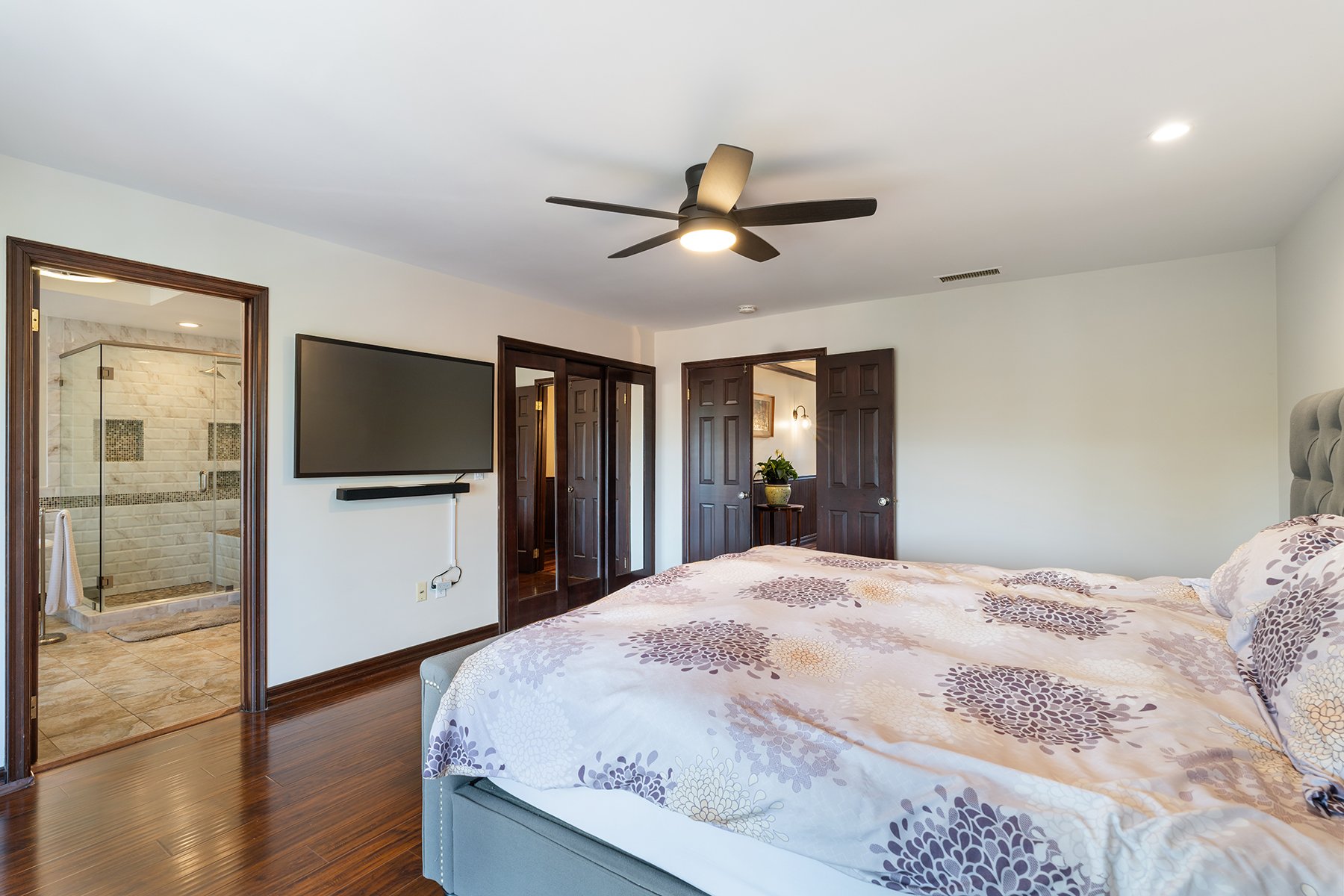 A bedroom with a bed in the foreground, a ceiling fan, a wall-mounted TV, and a bathroom with a glass shower door visible through an open door.
