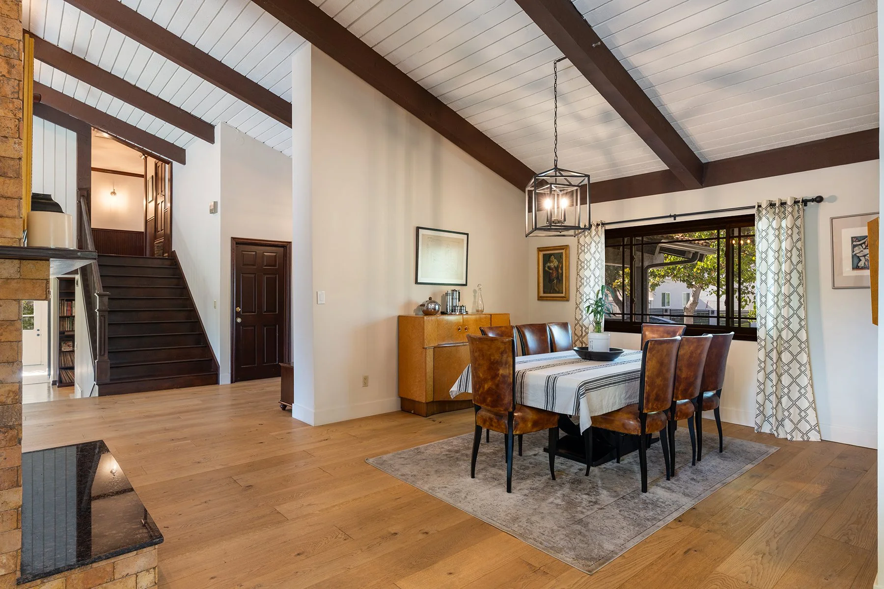 A dining room with a wooden table and six brown leather chairs, a window with curtains, a sideboard with decor items, and a chandelier hanging from a ceiling with exposed dark wood beams.