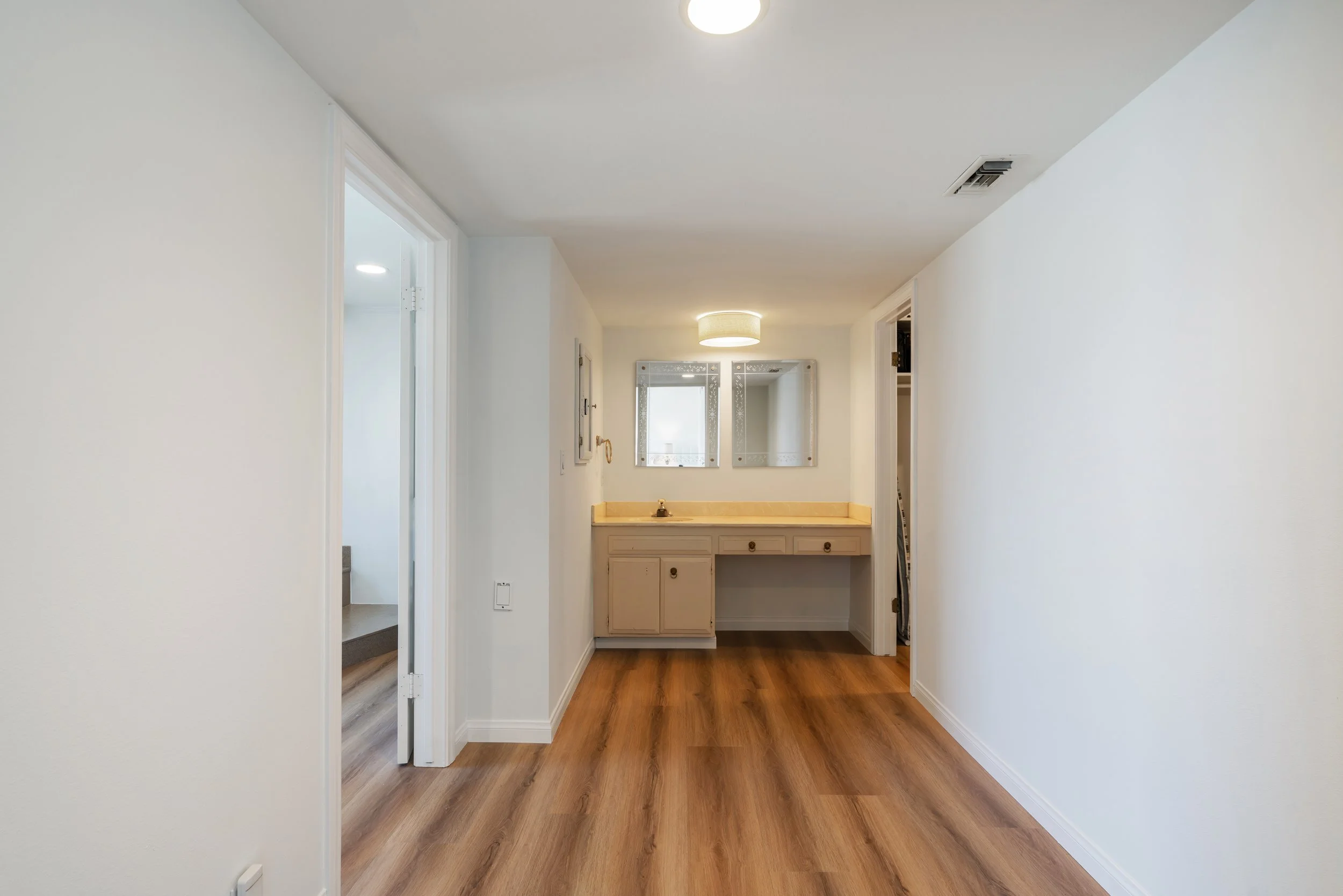 Empty room with wooden flooring, small vanity with a sink, two mirrors above, and a ceiling light fixture.
