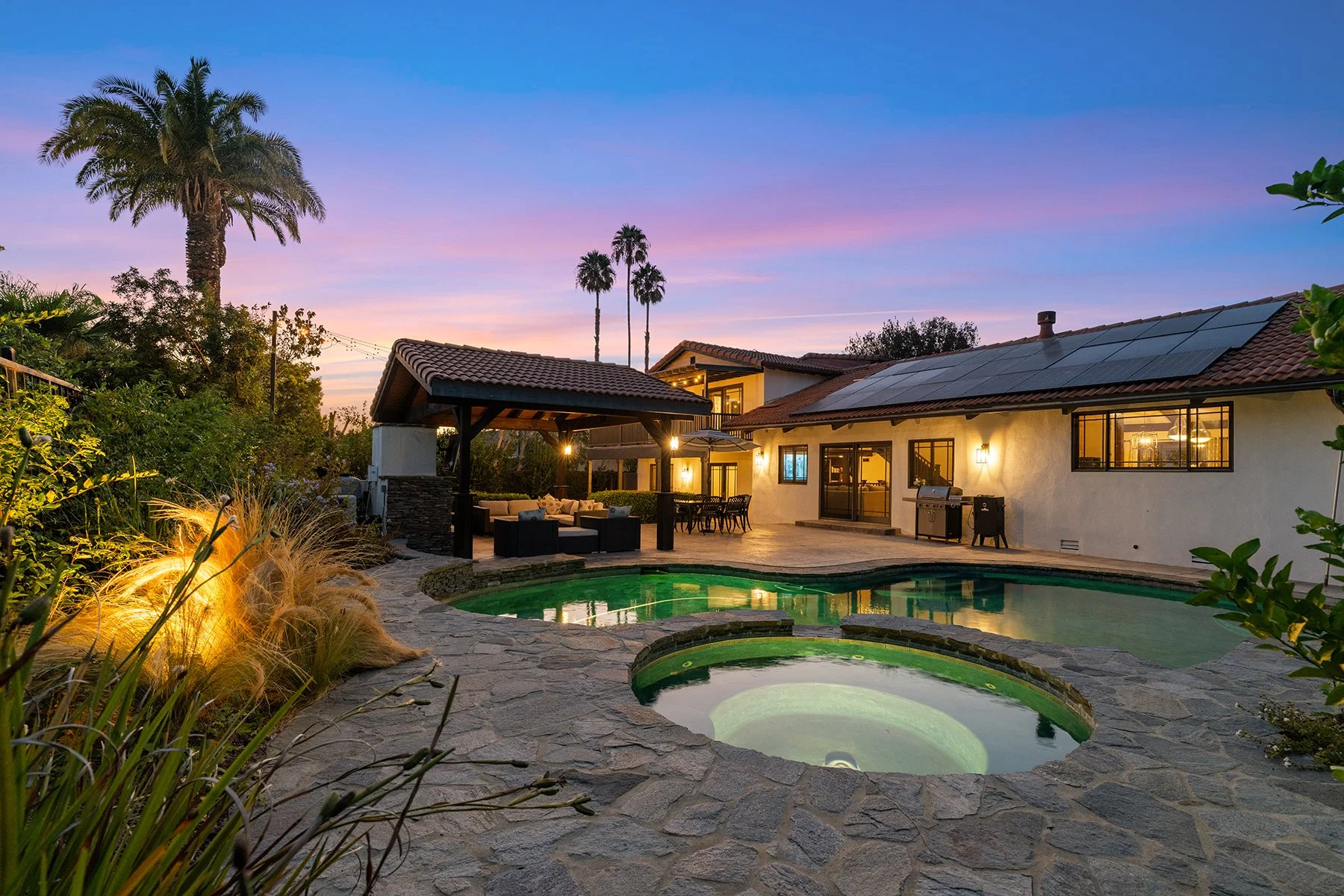 A backyard with a swimming pool and hot tub under the evening sky, surrounded by outdoor furniture, palm trees, and warm exterior lighting. Represented by Phil Missig and the Missig Team.