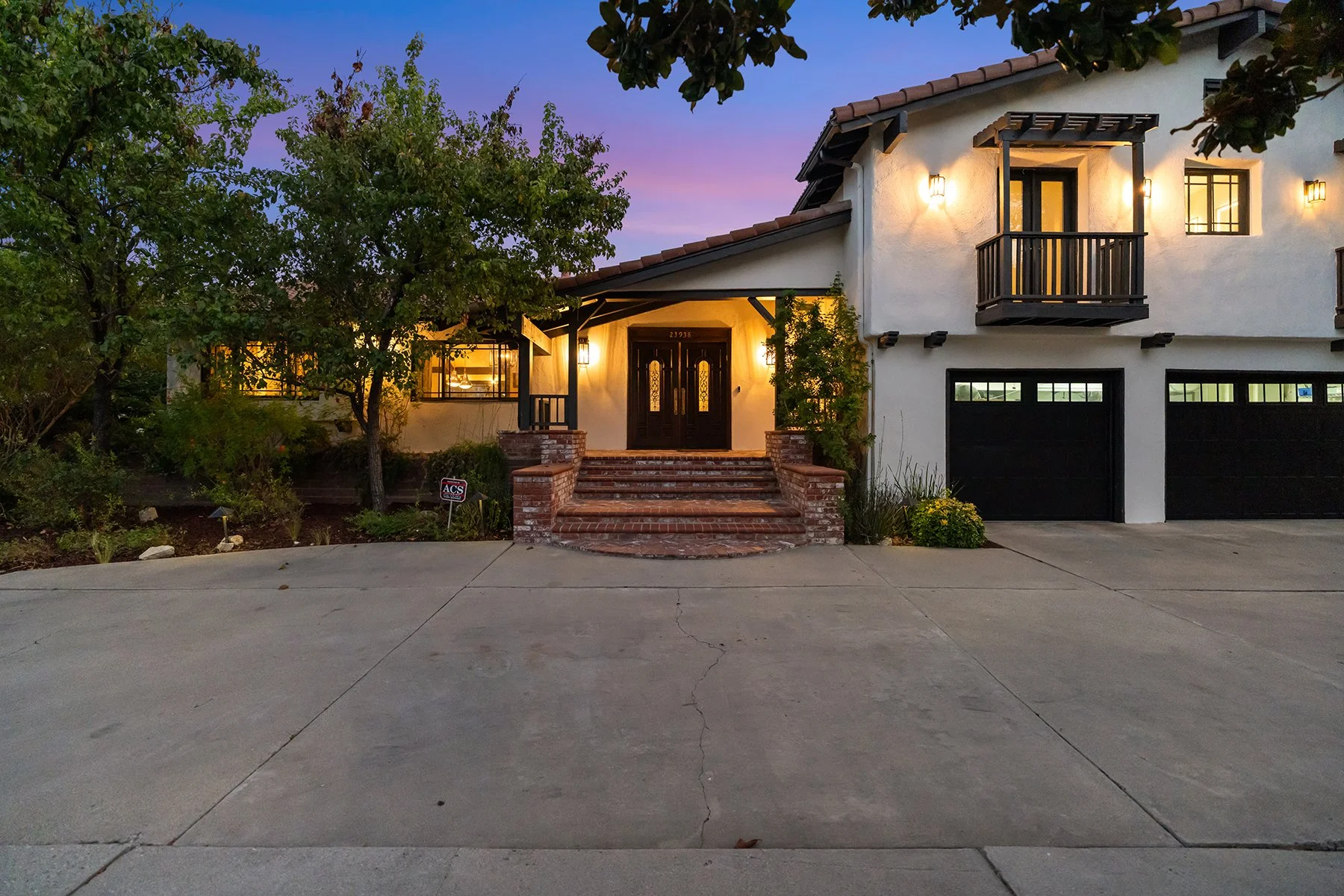 A two-story house with a white stucco exterior and dark brown trim, illuminated by exterior lights at dusk. The house has a small balcony above the front door, brick steps leading to the entrance, and a two-car garage with black doors. There are trees and bushes surrounding the house, and the sky is a gradient of purple and blue.