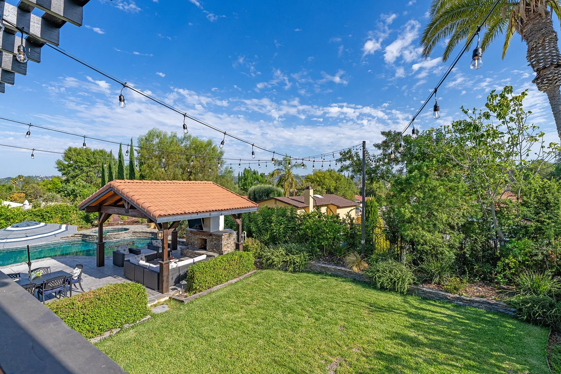A backyard with a swimming pool covered with a striped pool cover, outdoor furniture, a gazebo with a tiled roof, surrounded by lush green plants and trees, clear blue sky with clouds, string lights hanging above.