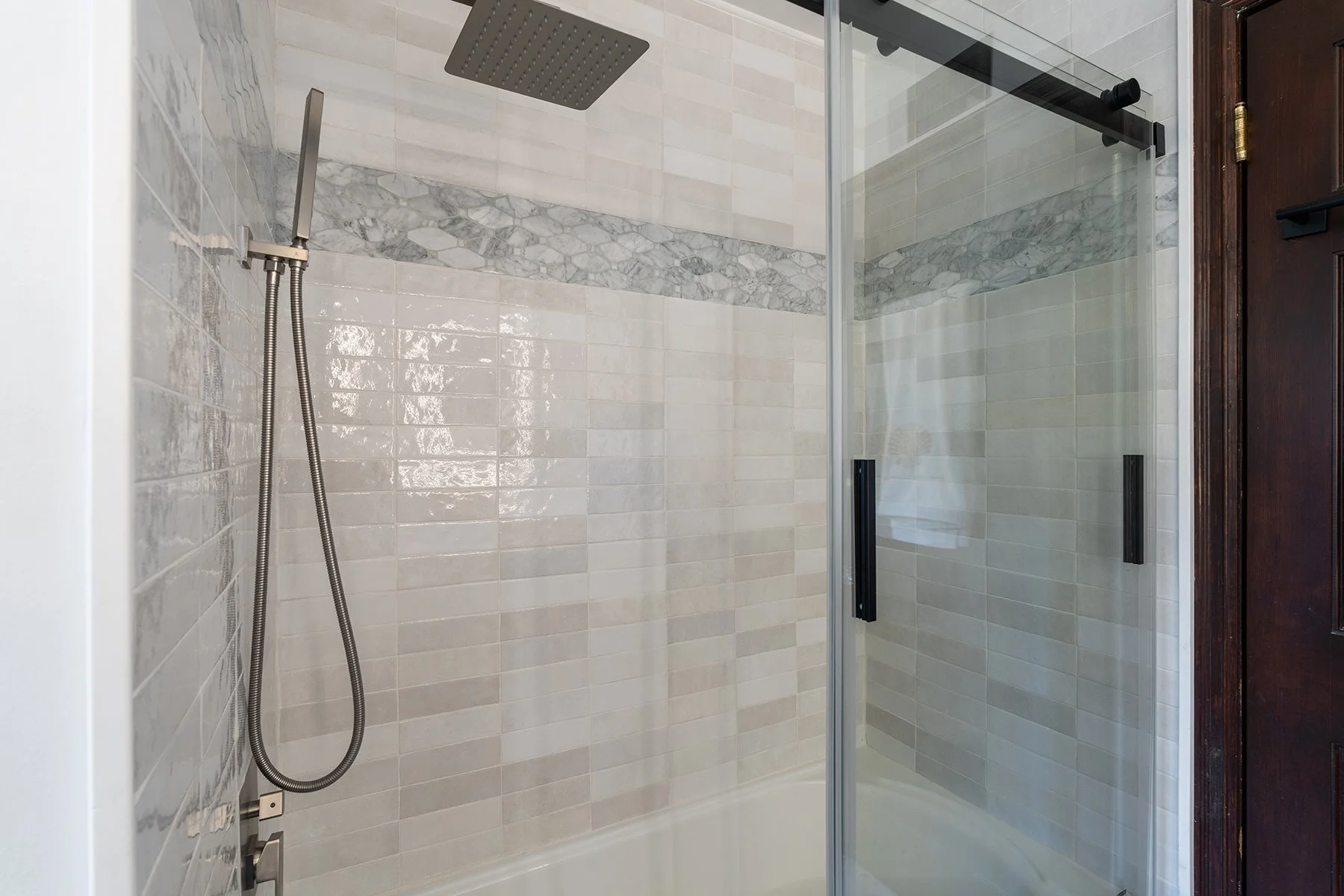 Modern walk-in shower with beige and marble tiles, a rainfall showerhead, a handheld shower, and a glass door with black handles.